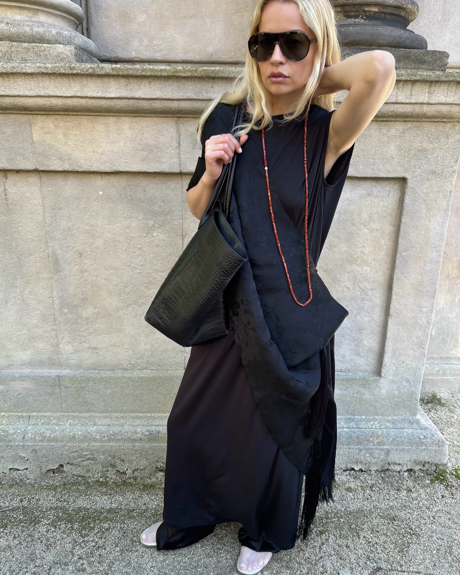 An image of an influencer standing in front a building wearing a draped navy scarf top, wide-leg black trousers, white mesh flats, black aviator sunglasses and a long red beaded necklace.