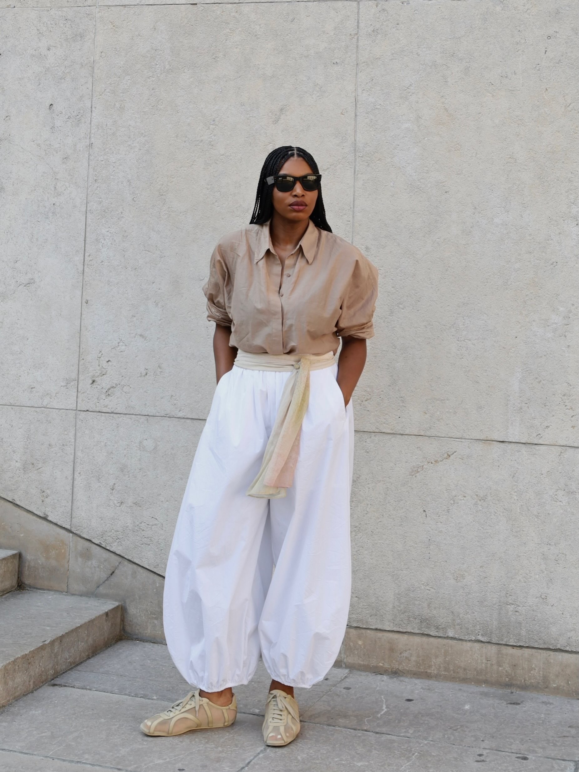 An image of an influencer wearing black sunglasses, a beige shirt, white cotton poplin balloon trousers and beige trainers standing in front of stone wall