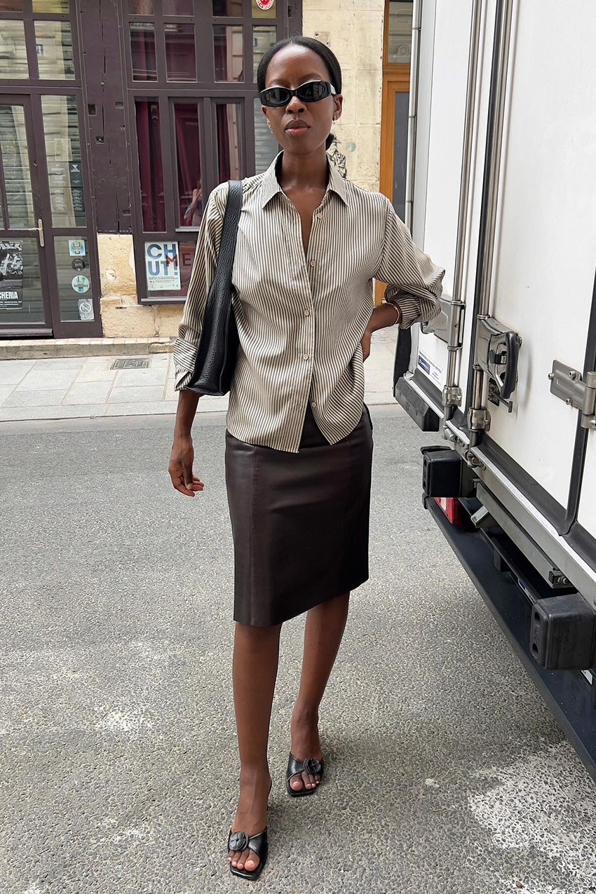 A woman wearing a striped button-down, brown leather skirt, and black sandals.