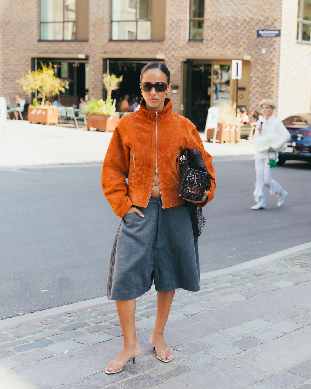Woman wears orange suede jacket, gray trouser shorts, thong sandals, and a handbag.