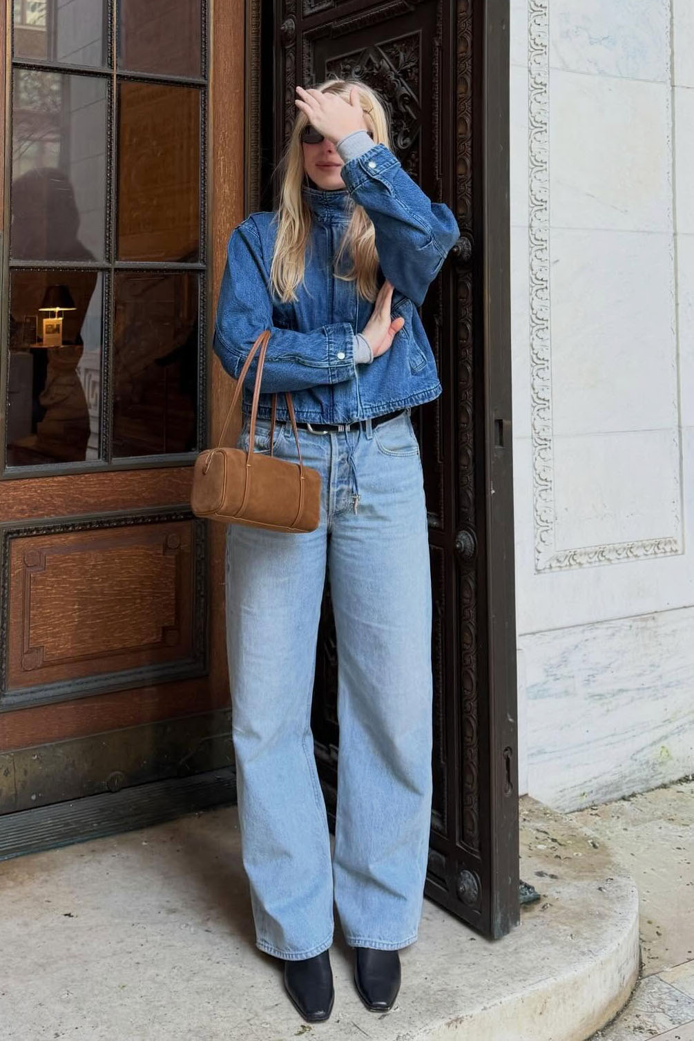 A woman wearing a denim jacket, blue barrel jeans, black boots, and a brown bag.