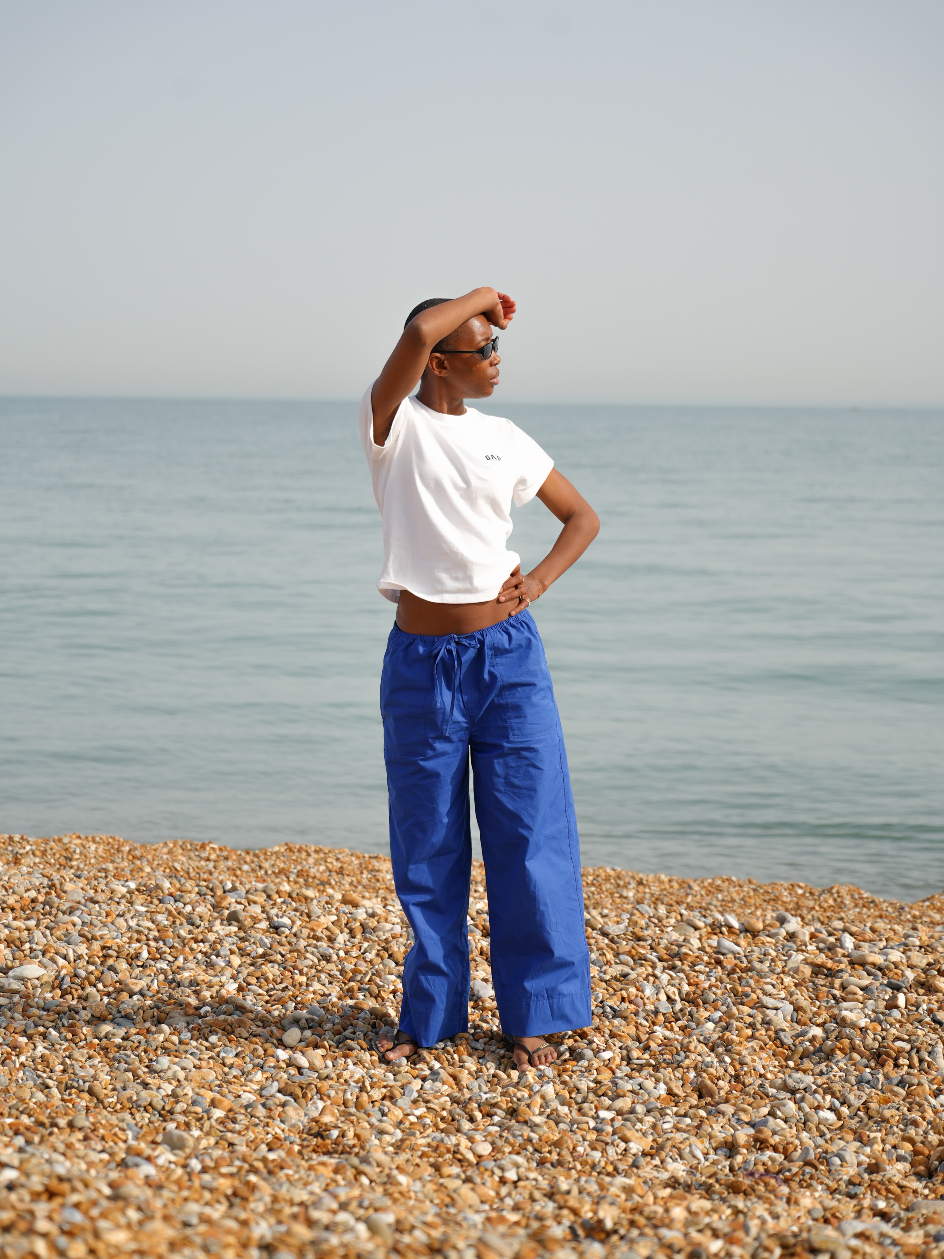 An image of an influencer standing on a stony beach wearing black sunglasses, a white T-shirt and cobalt blue cotton poplin trousers