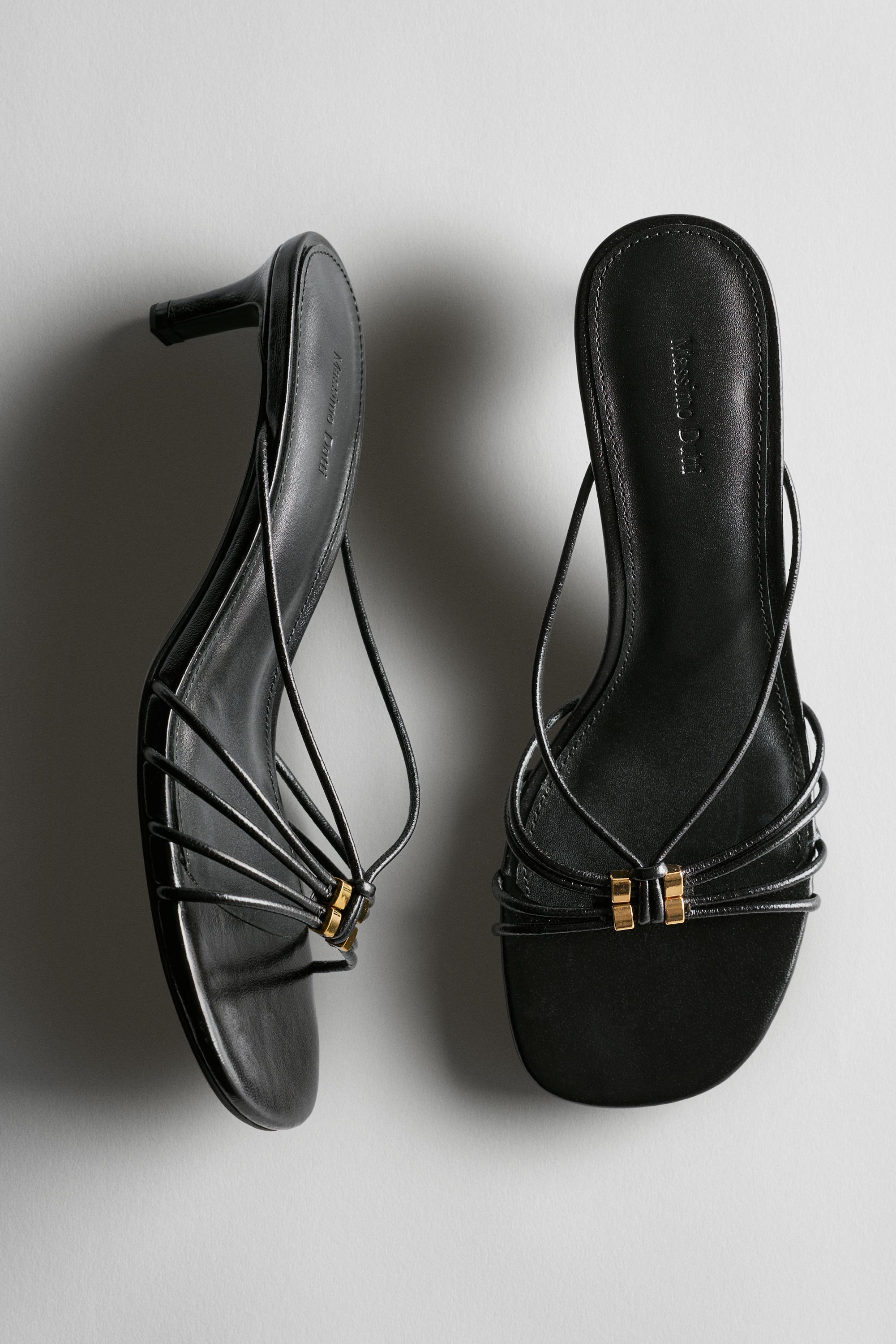 A photo of a pair of black strappy heeled sandals with gold embellishments on the front