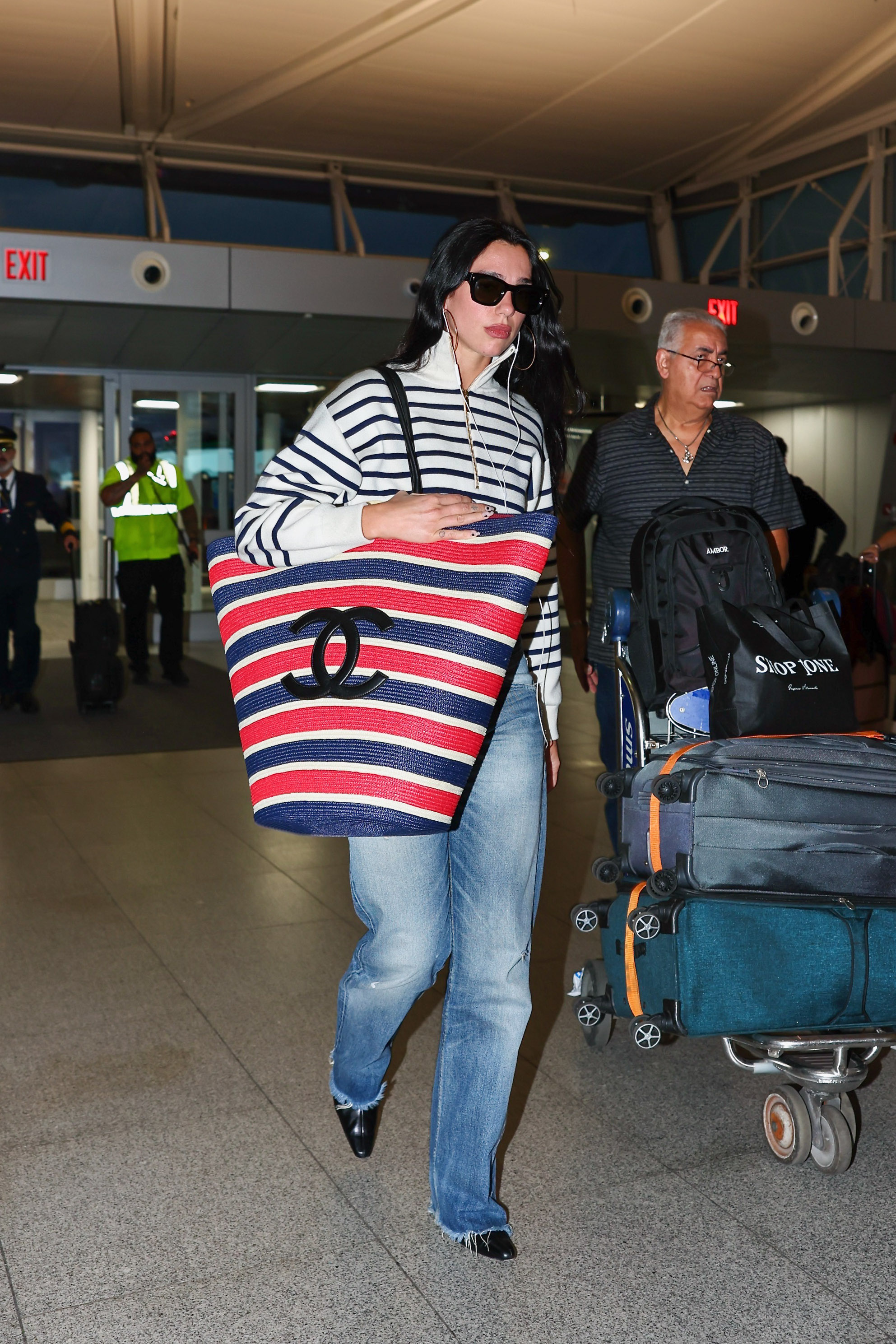 Dua Lipa walks through the airport carrying an oversized Chanel tote bag wearing a striped knit with jeans and black pointed-toe heels.