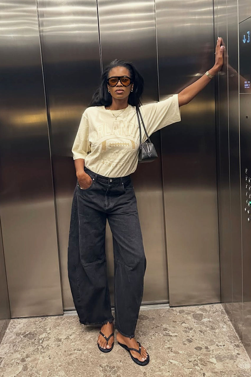 A woman wearing a white T-shirt, black barrel jeans, and black flip-flops.