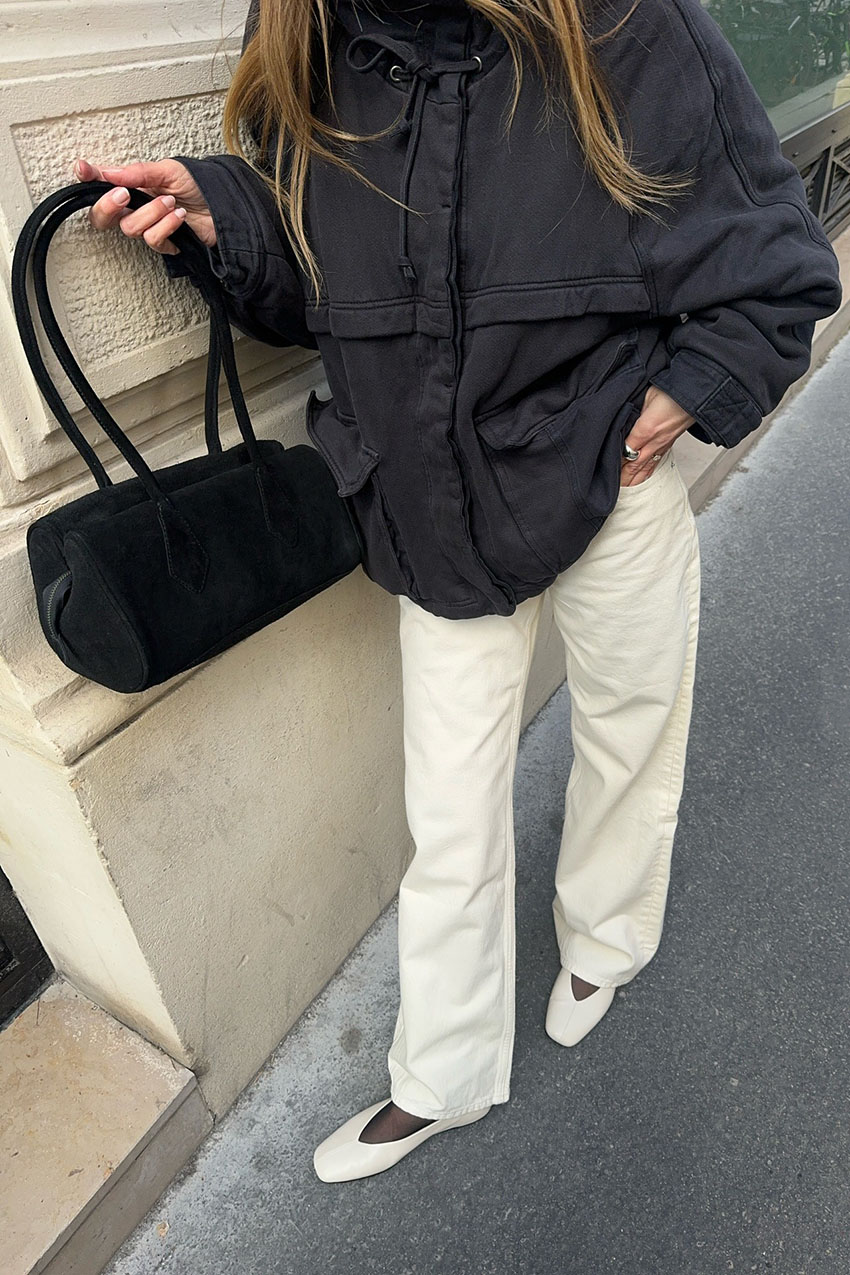a woman wearing a black jacket, white pants, white flats, and black east-west bag