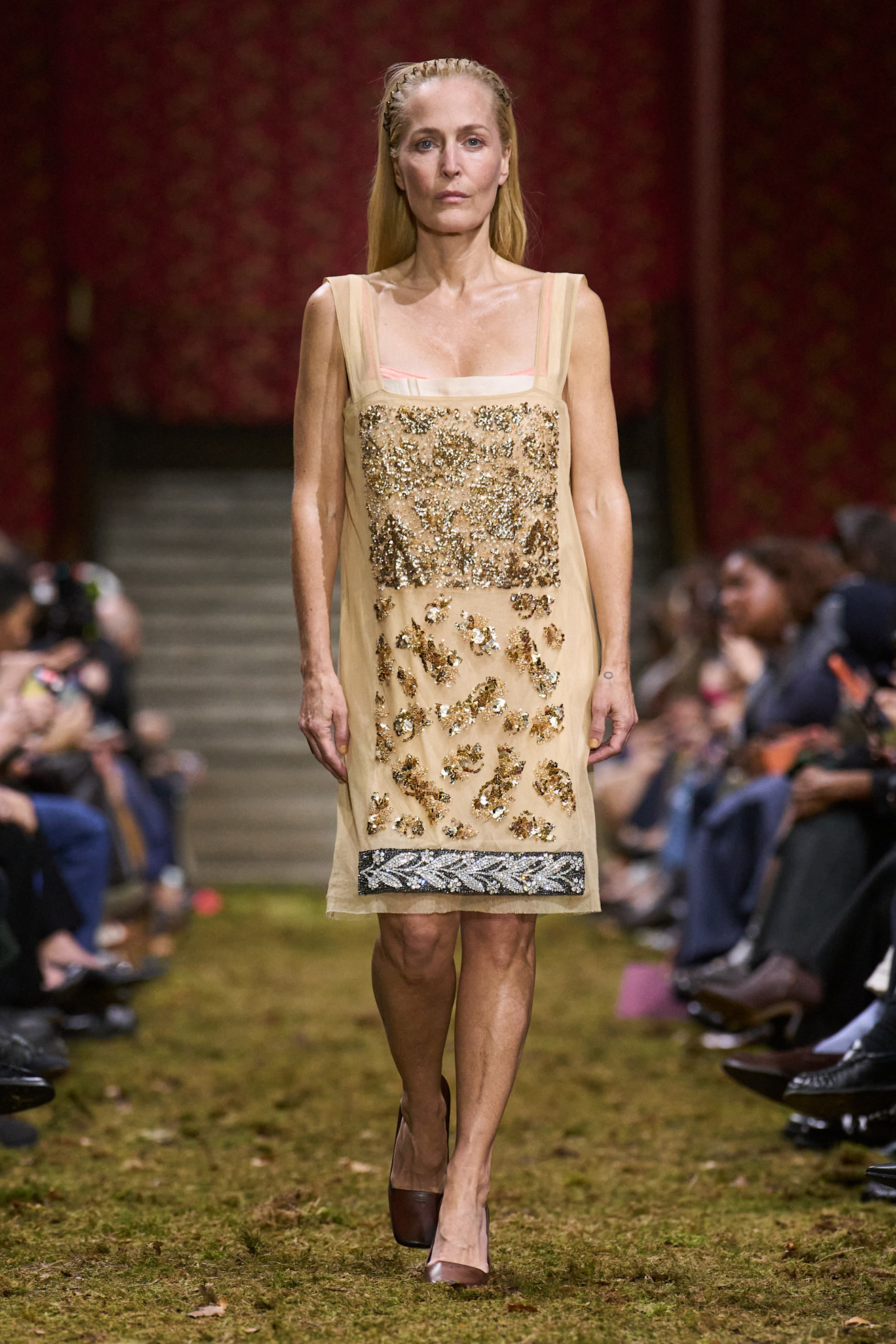 An image of actor Gillian Anderson wearing a beige dress with gold and silver beaded embellishments walking down a moss-covered runway for the Miu Miu AW26 show