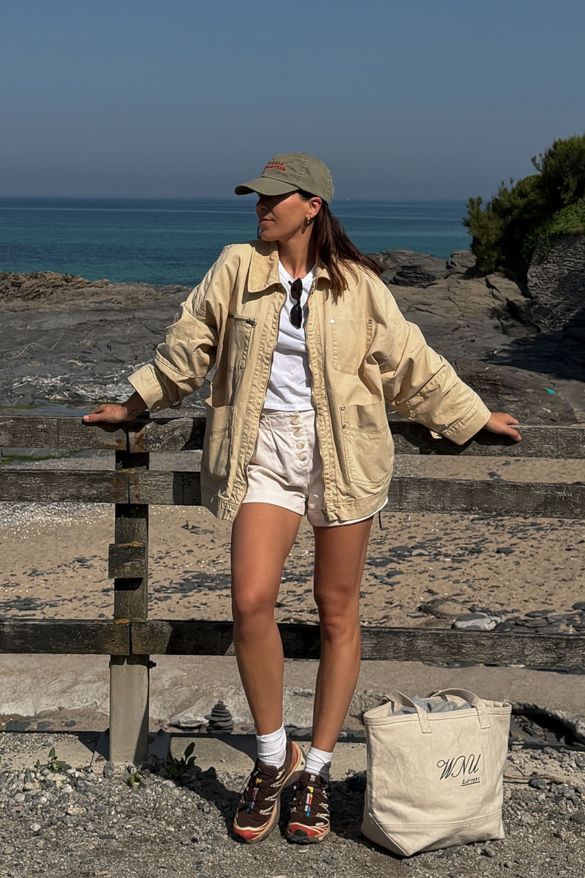 a woman wearing a baseball cap, tank jacket, white tee, shorts, and chunky sneakers