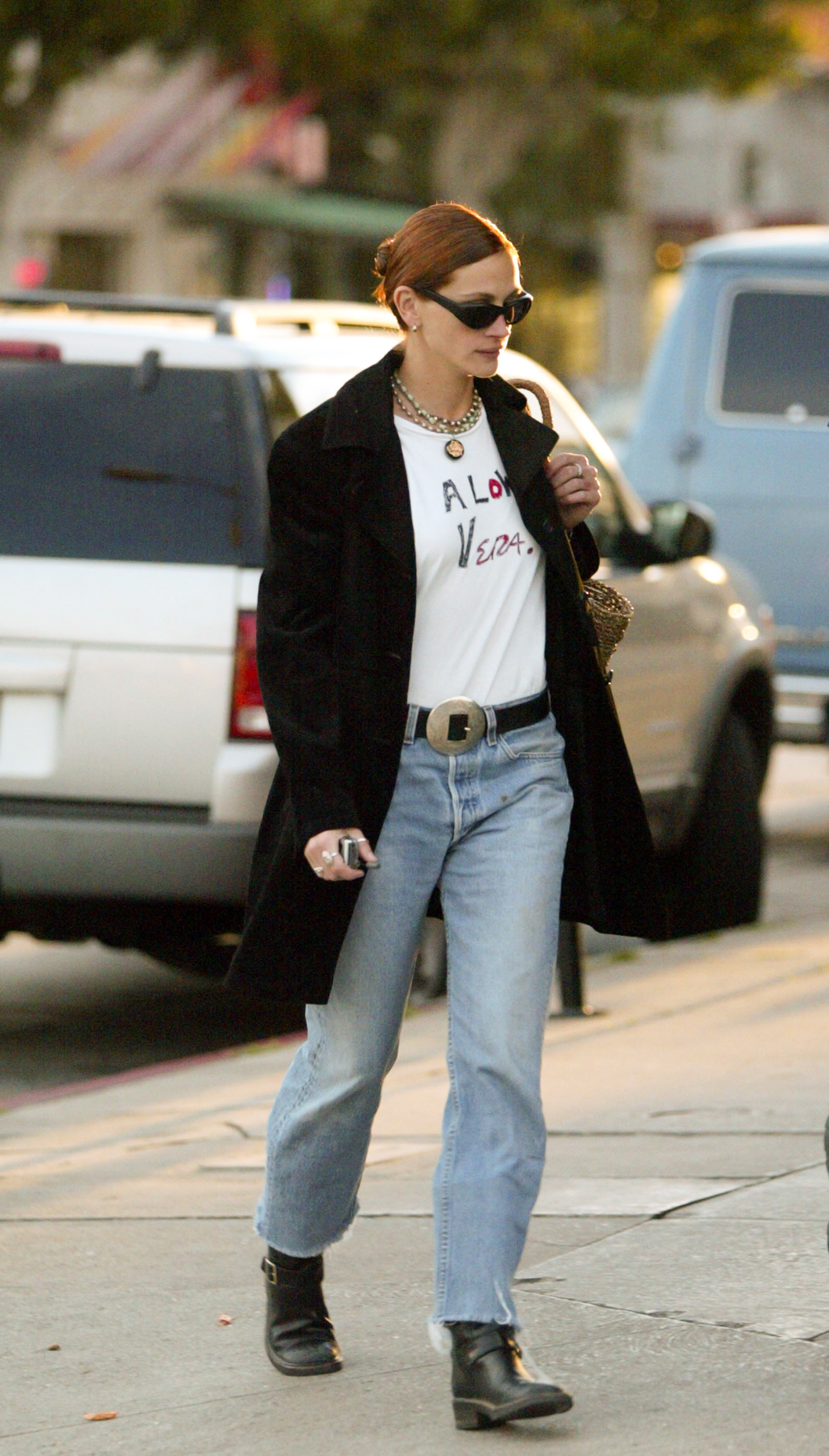 An image of actor Julia Roberts from the '90s walking outside wearing a black coat, a graphic T-shirt that says 