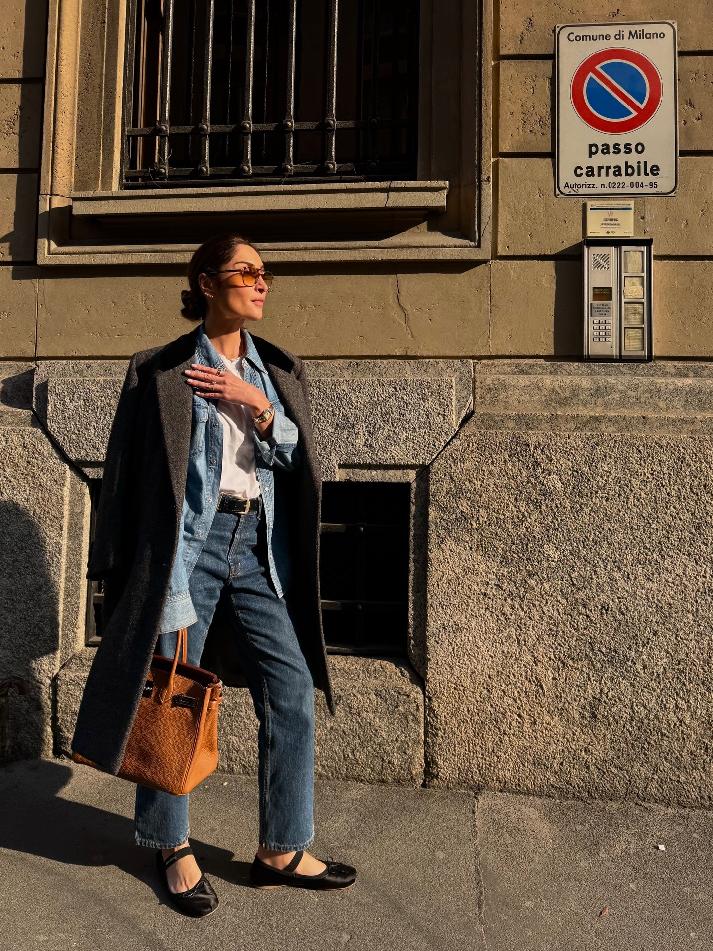 French woman wearing a travel outfit that includes a coat over a denim shirt and a white tee with jeans and black ballet flats