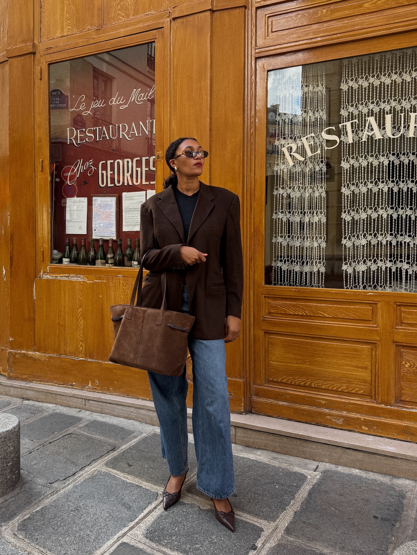 An image of an influencer wearing a brown blazer and blue jeans, holding a brown suede tote and wearing brown heels in Paris