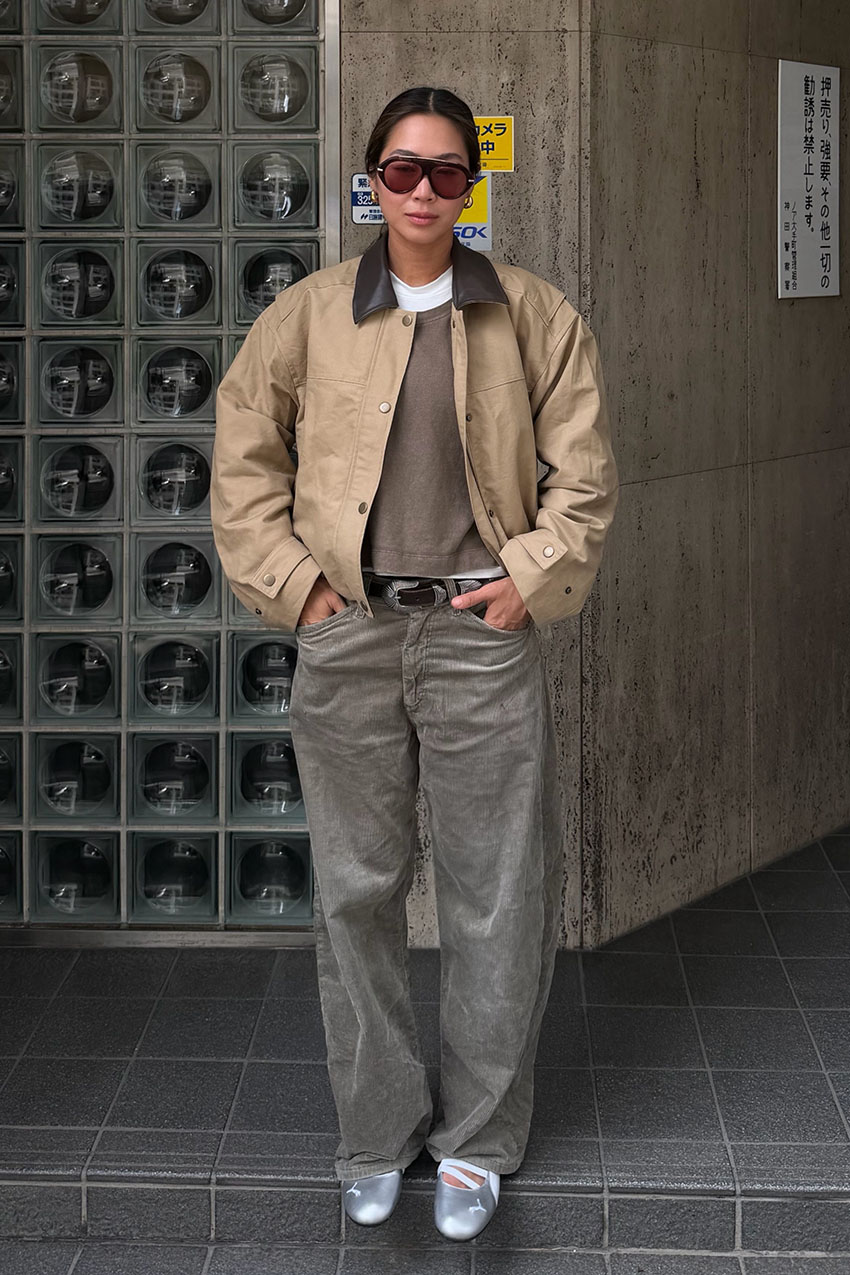 A woman wearing a contrast-collar jacket, brown sweater, gray barrel jeans, and silver flats.