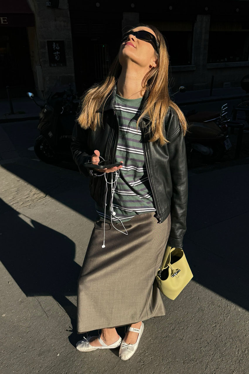 a woman wearing a black leather jacket, striped shirt, khaki skirt, and satin flats