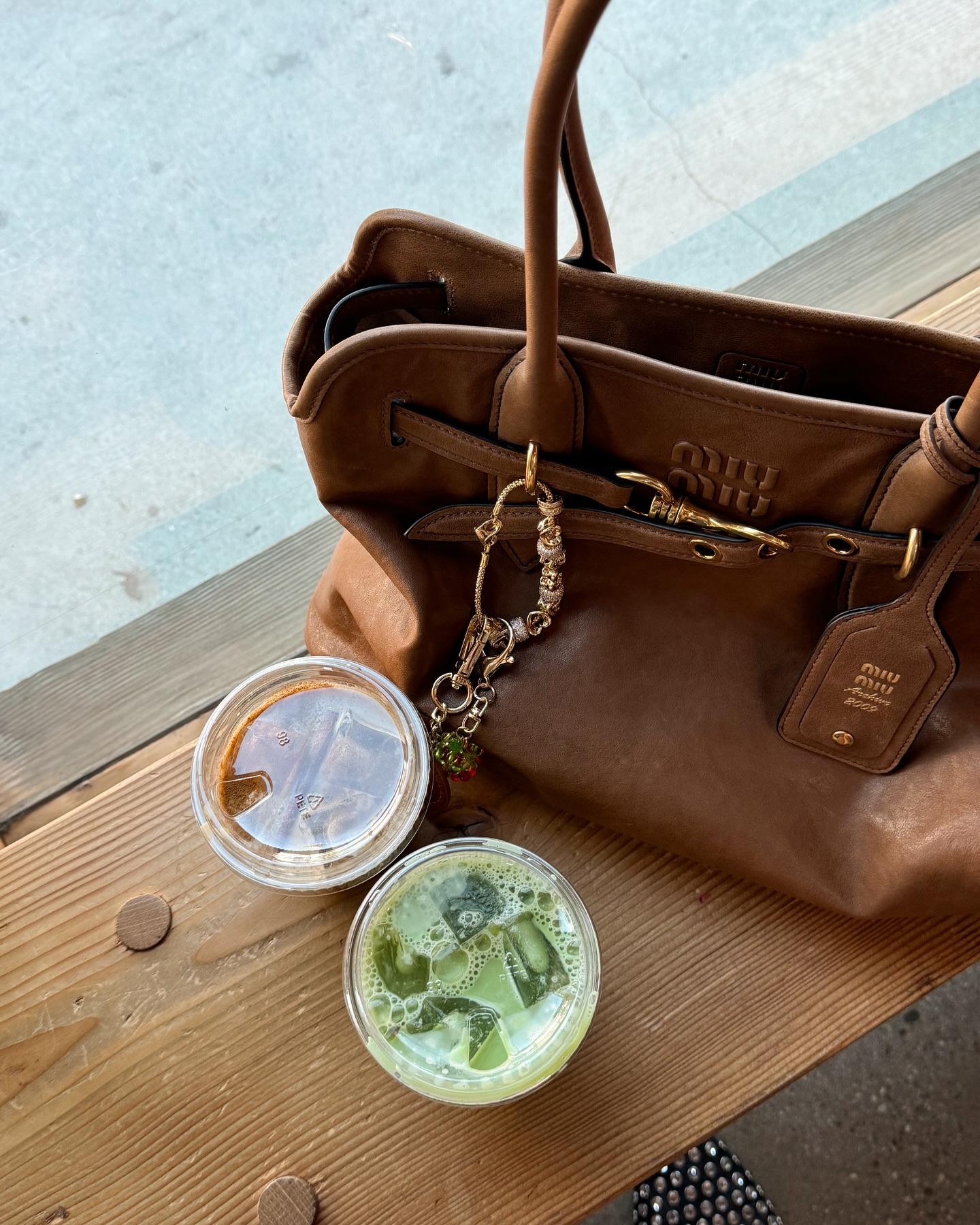An image of a brown Miu Miu bag with gold keychains on a wooden table next to an iced coffee and match