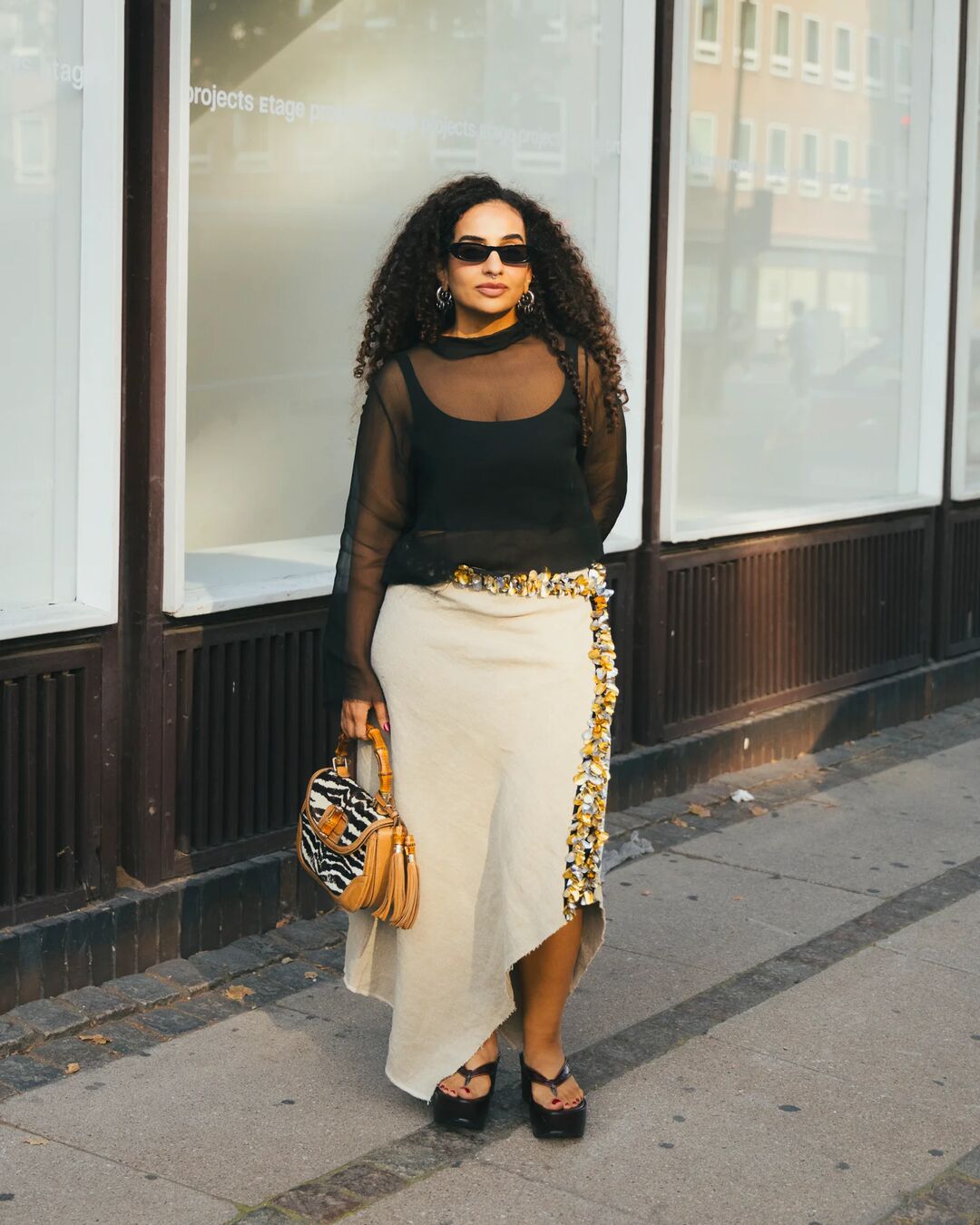 Woman wears long sleeve sheer top, asymmetrical beaded handbags, platform flip flops, and zebra printed handbag.