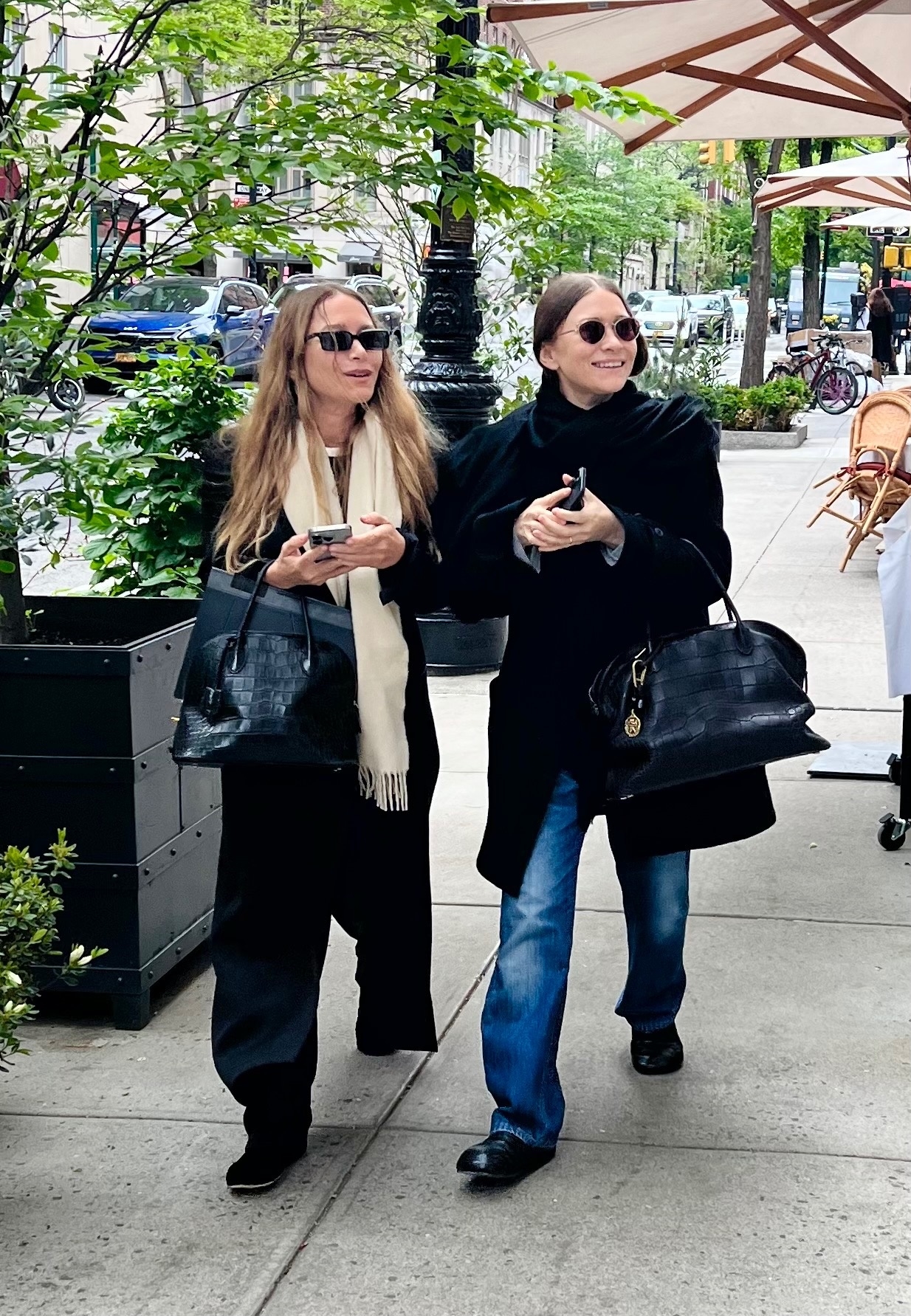 An image of Mary-Kate and Ashley Olsen in New York. Mary-Kate wears a black blazer with black trousers and slippers. Ashley wears a black blazer with jeans and loafers.