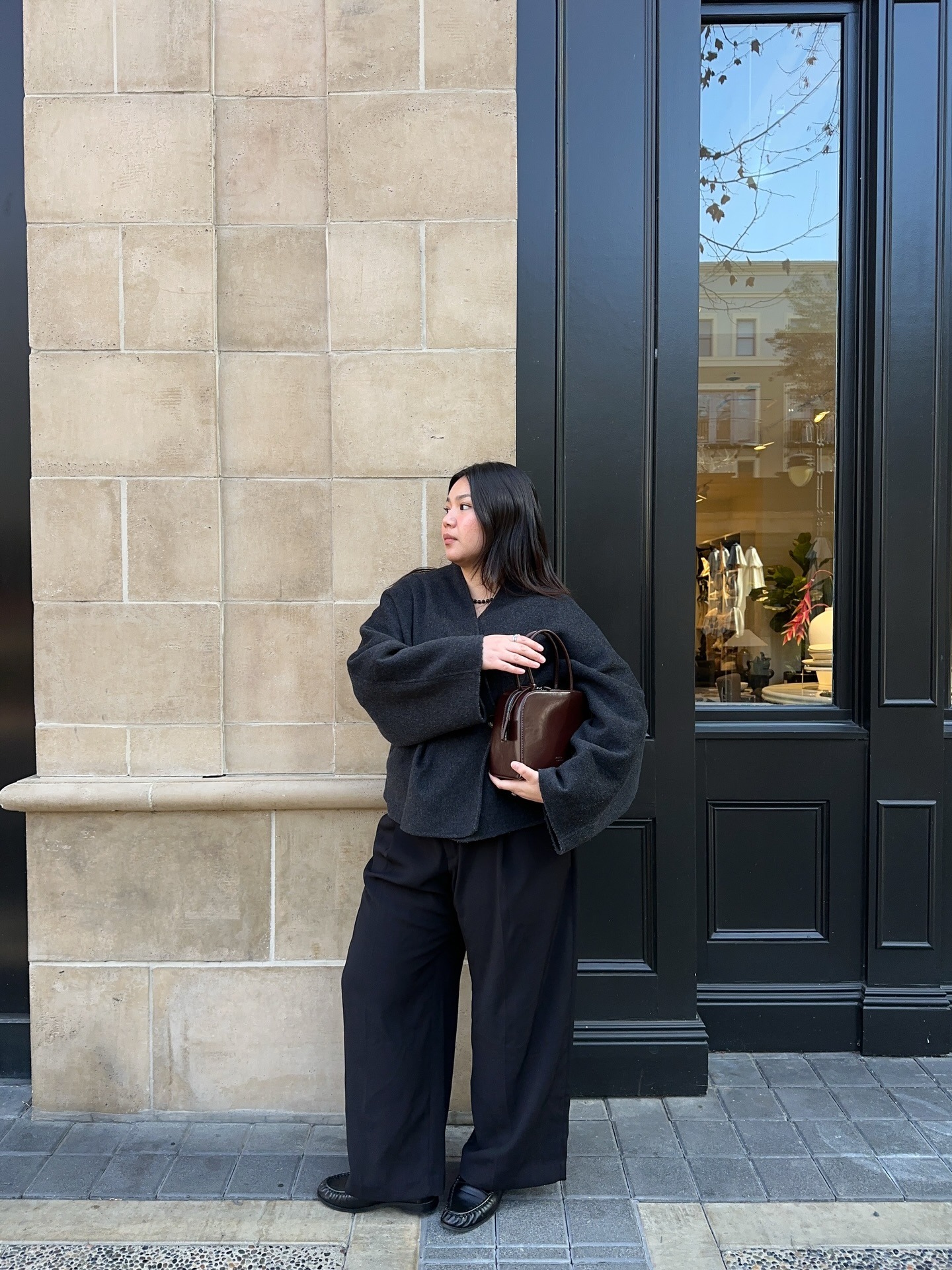 Marina wears black jacket/coat with black trousers and black loafers while carrying a brown handbag.