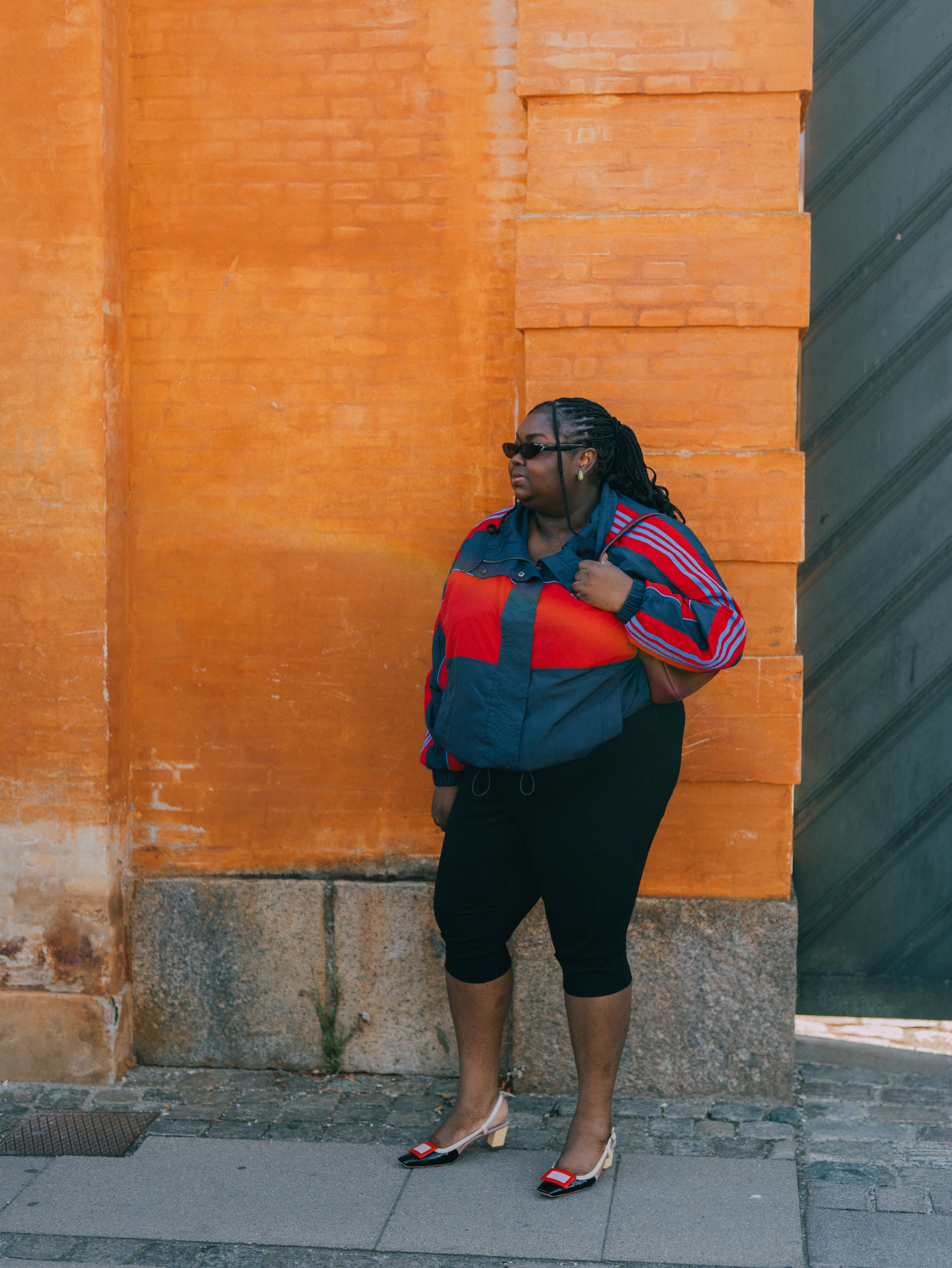 An image of an influencer standing in front of an orange brick wall wearing a teal and red windbreaker with black capri leggings and beige, black and red heels