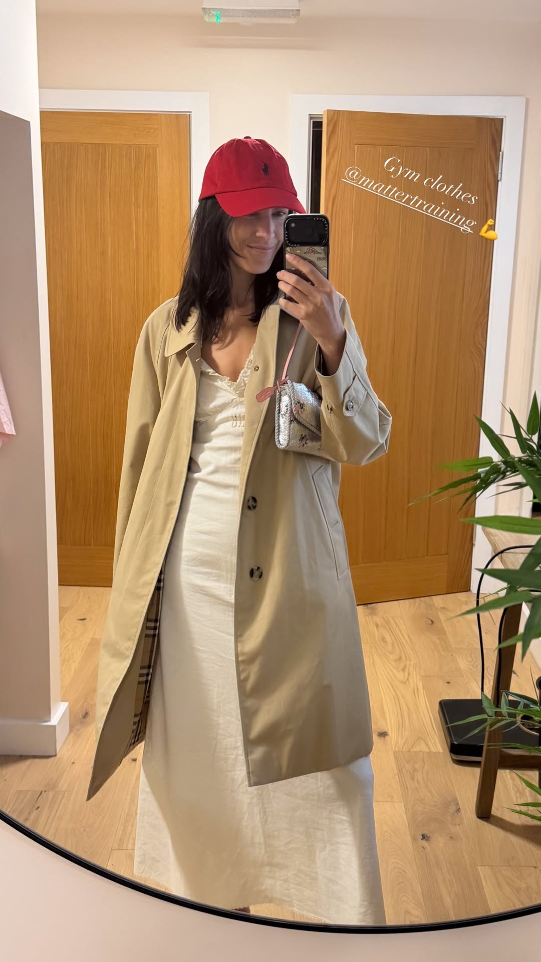 Alexa Chung takes a mirror selfie wearing a white cotton dress with a beige trench coat and a red cap.