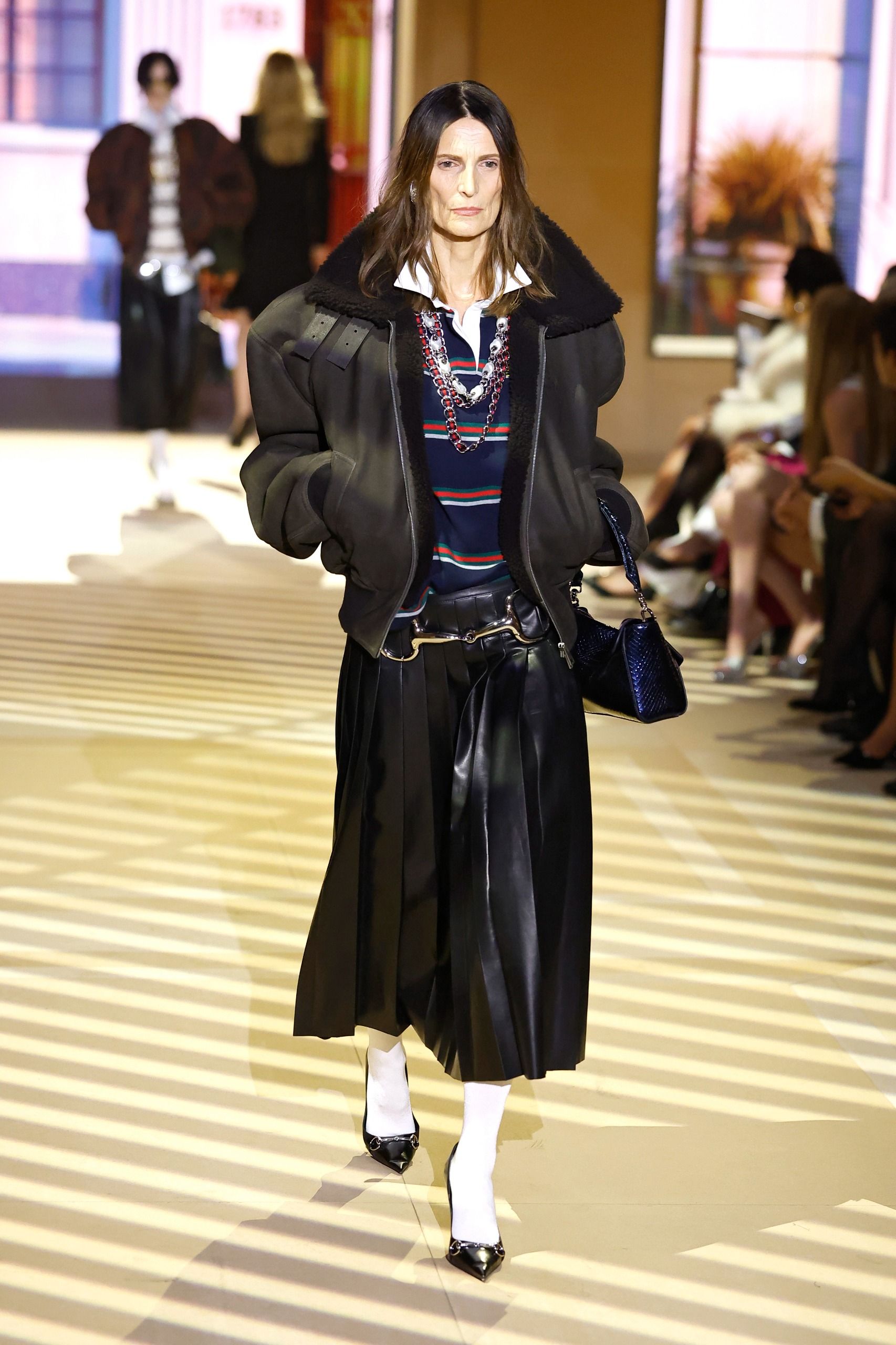 Model walks the Gucci SS27 Runway wearing layered chunky necklaces with a leather pleated skirt, a polo knit and a leather jacket.