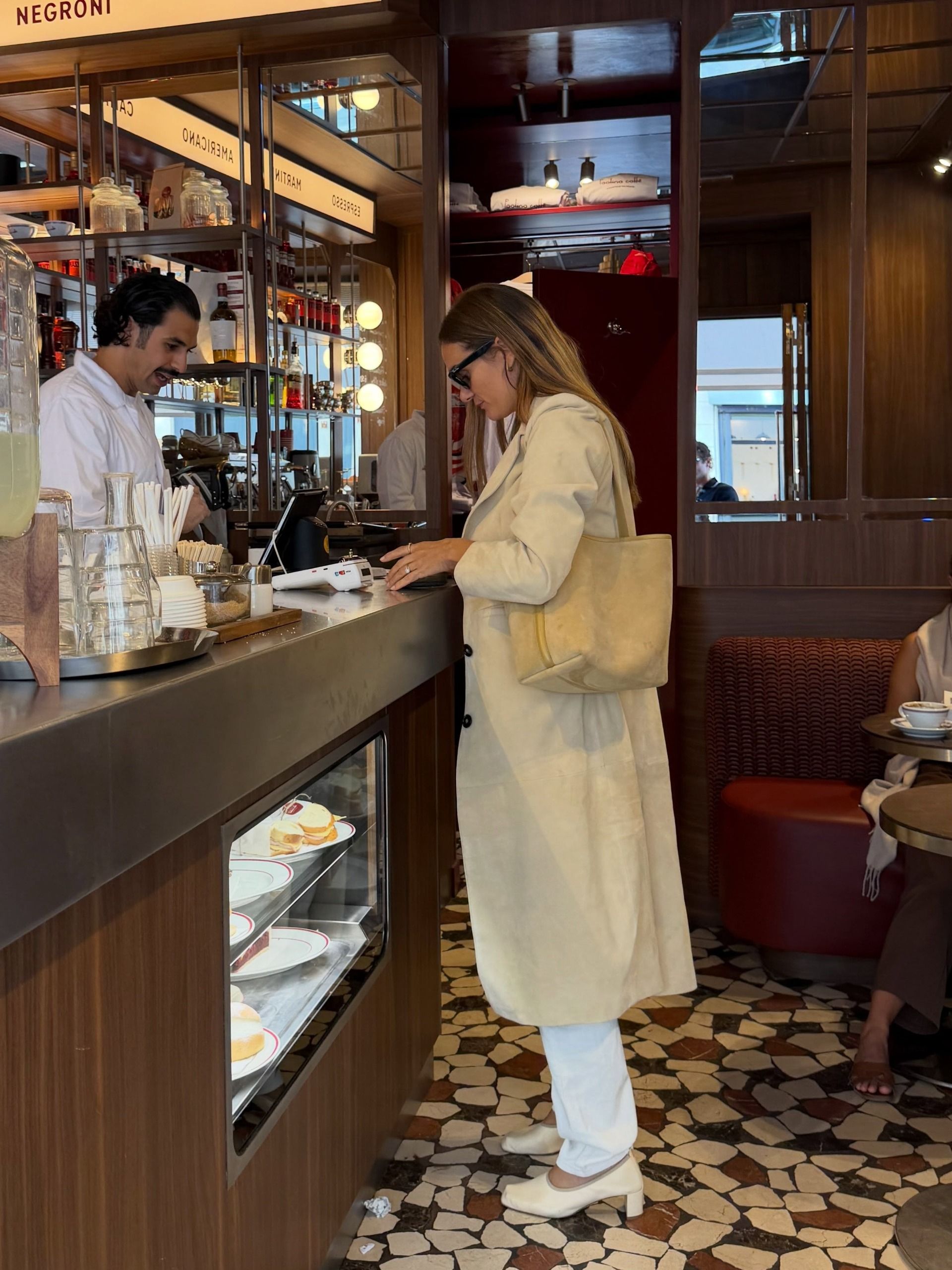 An influencer standing in a cafe in Paris wearing a trench coat with white jeans and cream-coloured ballerina pumps, carrying a suede tote on her shoulder
