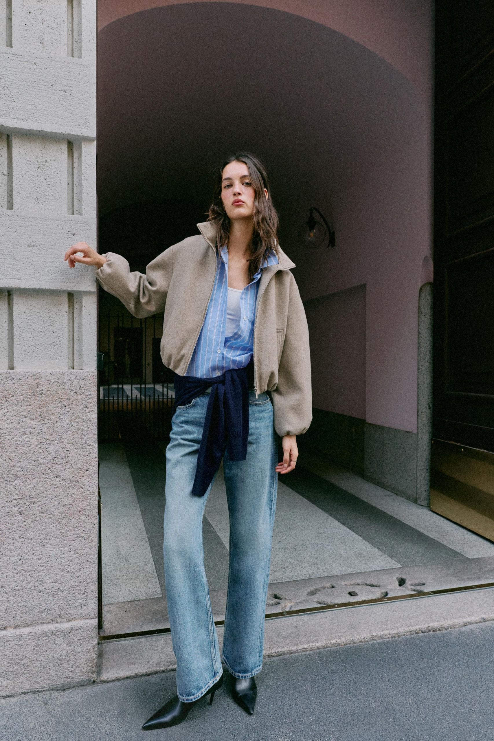 Woman wears blue button down with tan bomber and blue jeans.