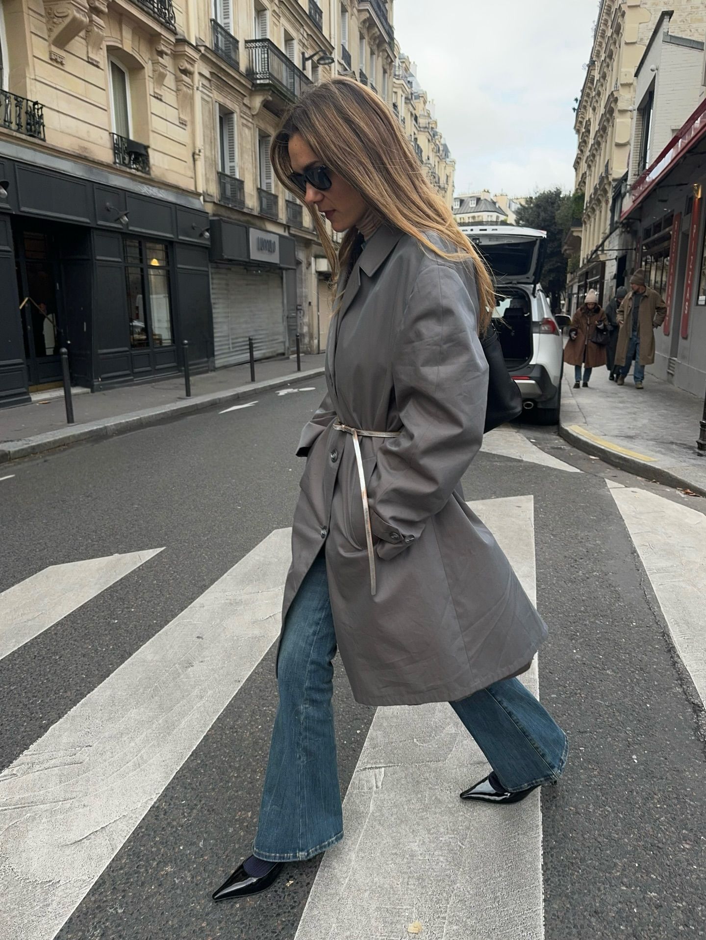 An image of an influencer in Paris wearing a grey trench coat with a silver belt with flare jeans and black pointed-toe patent leather heels