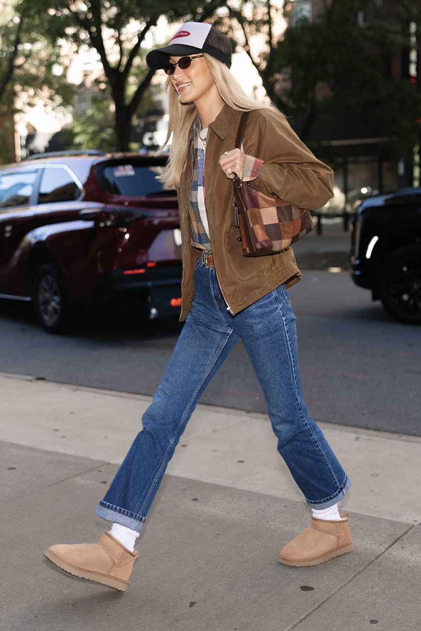 Bella Hadid wearing a brown collared jacket, plaid shirt over a T-shirt, and jeans with a baseball cap, Coach Alter/Ego Slouchy Shoulder Bag, and Ugg Classic Ultra Mini Boots with white socks