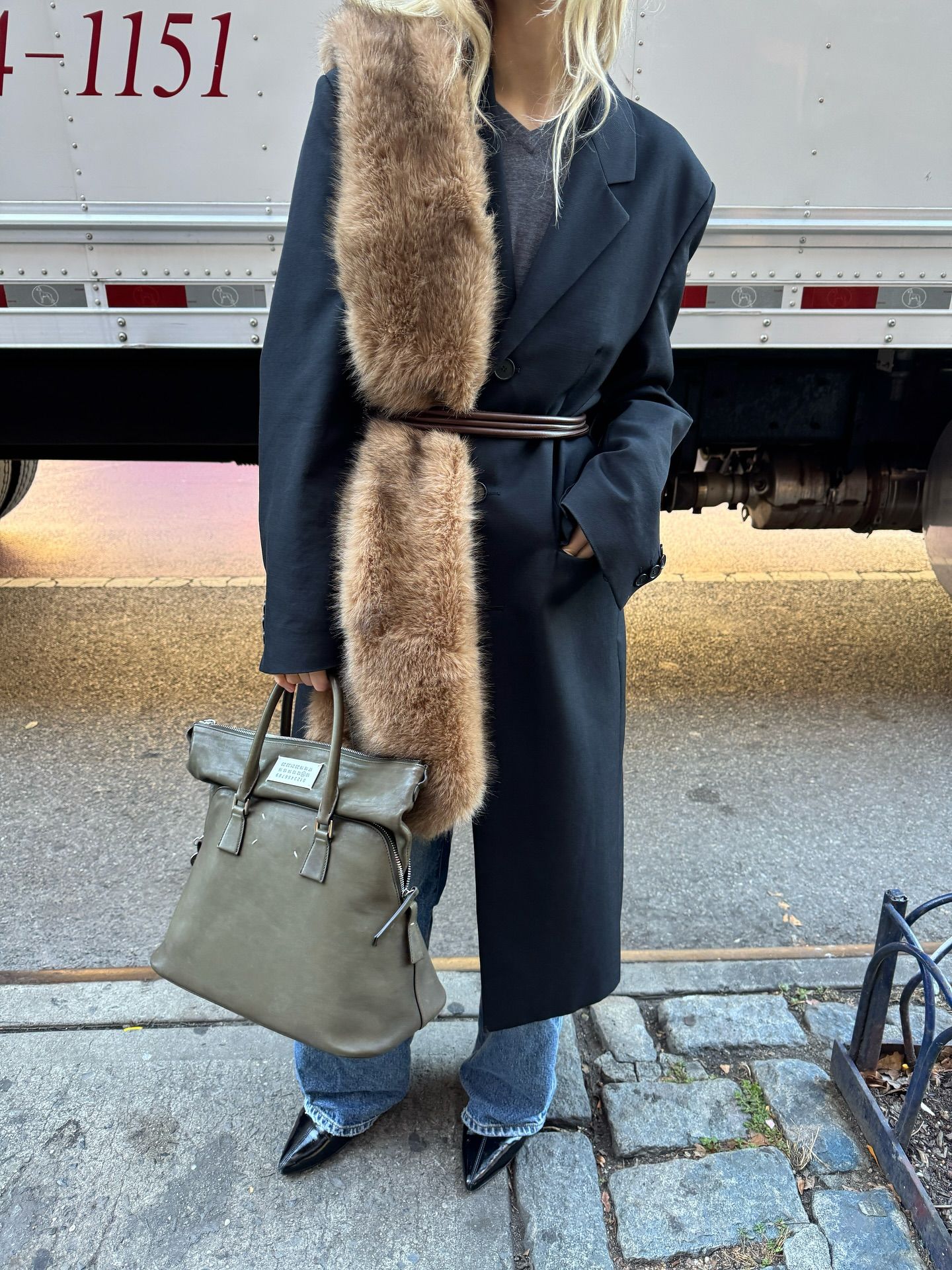 An image of an influencer in New York wearing a long black tailored coat with a fur scarf over top of jeans and pointed-toe shoes holding an olive green tote