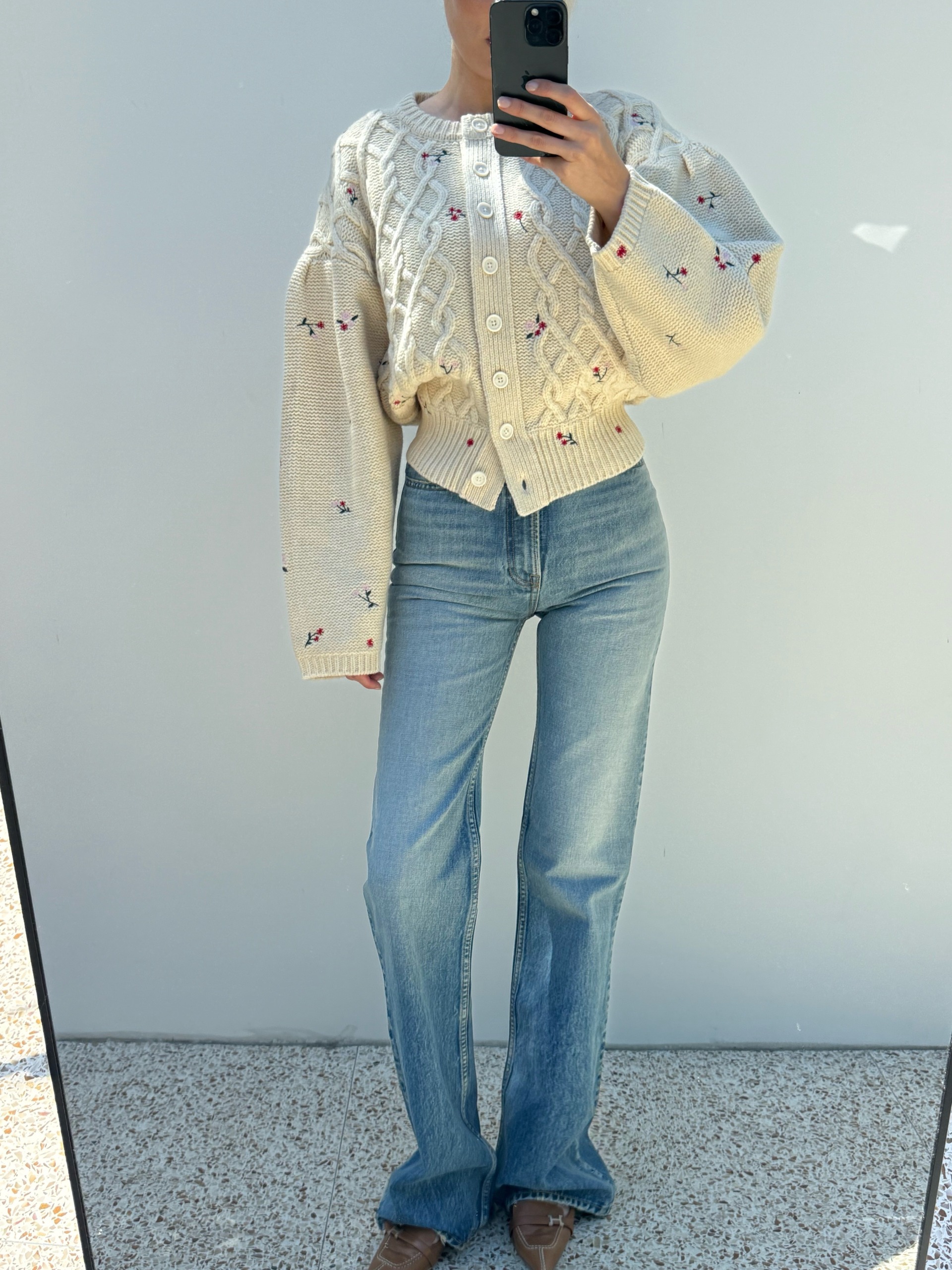 Model Elsa Hosk takes a mirror selfie wearing mid-wash bootcut jeans with brown loafers and a cream, printed cardigan.