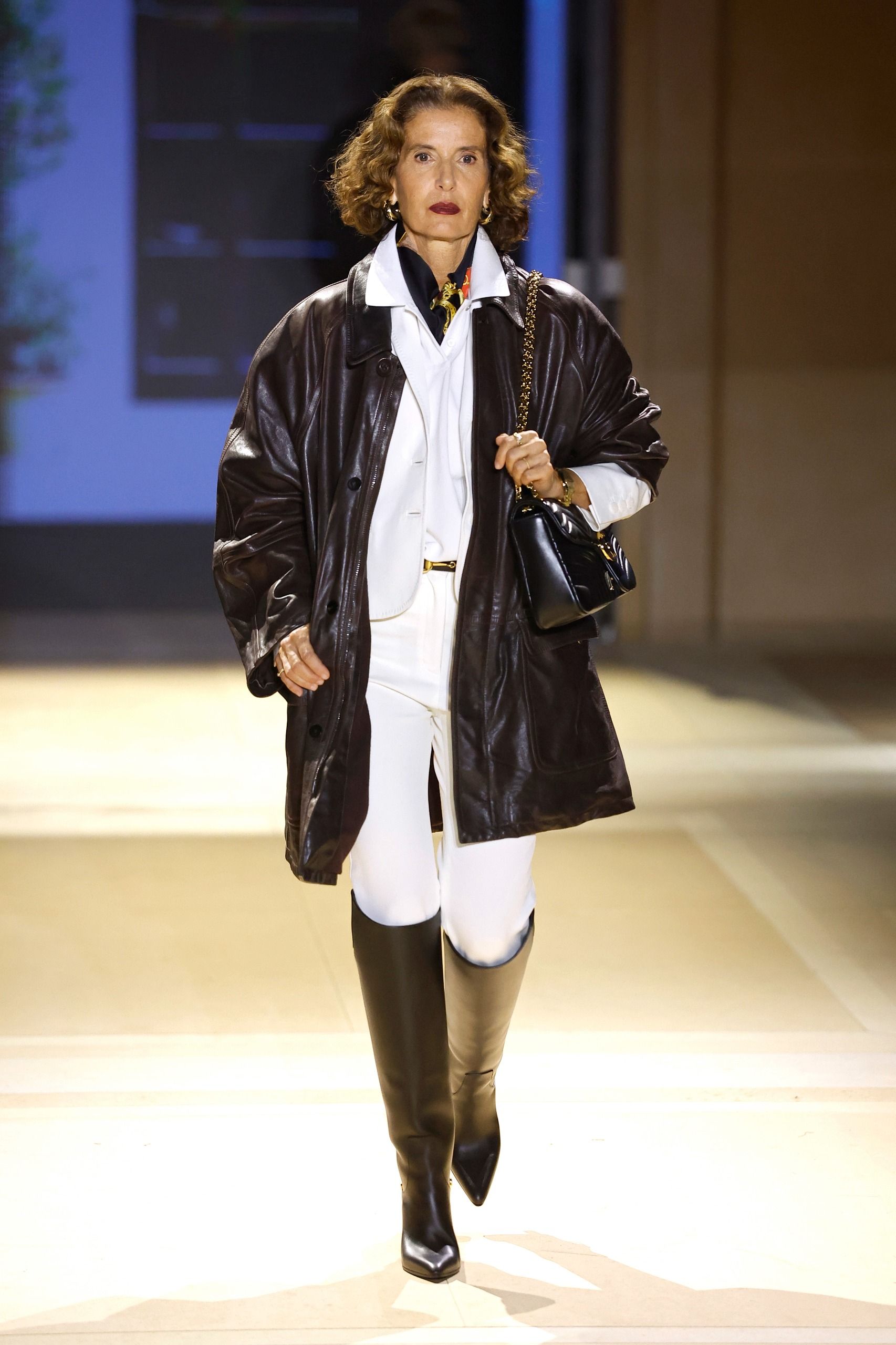 Model walks the Gucci SS27 Runway wearing a white collared shirt layered over a black leather jacket with white leggings and black knee-high boots.