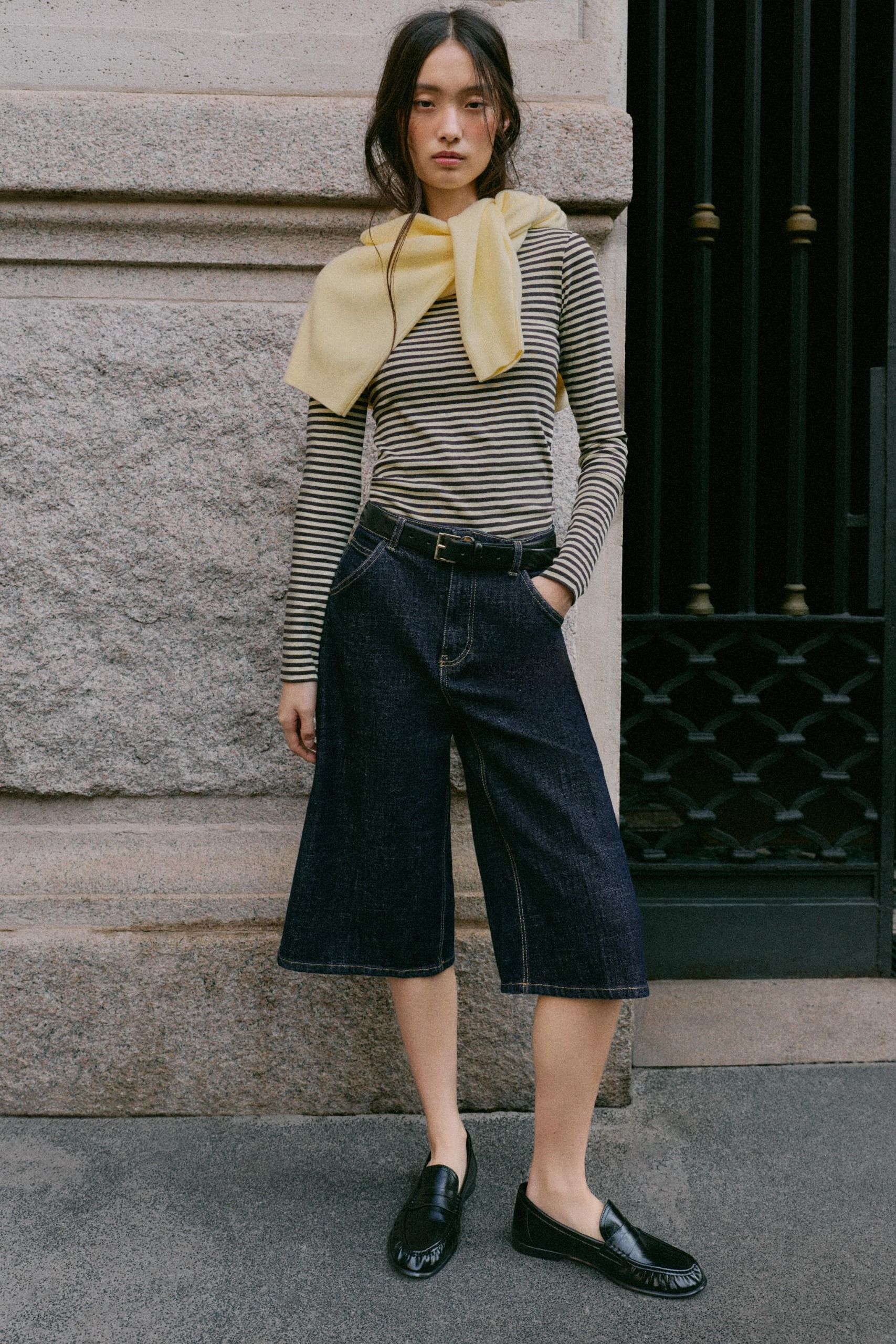 Woman wears black and white striped shirt, long denim short/culottes, and black loafers.