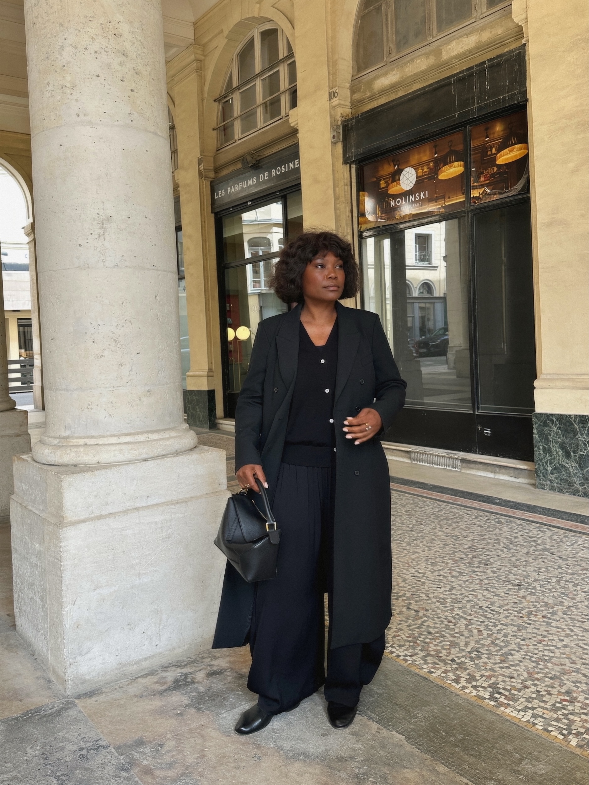 An image of an influencer in Paris wearing a black tailored coat, a black cardigan, navy balloon trousers, black ballet flats and holding a black leather handbag
