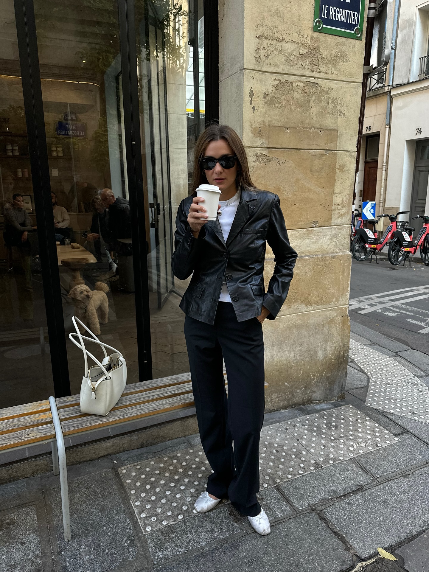 An image of an influencer in Paris wearing black sunglasses, a black leather blazer over a white T-shirt, black trousers and white satin ballet flats holding a coffee cup
