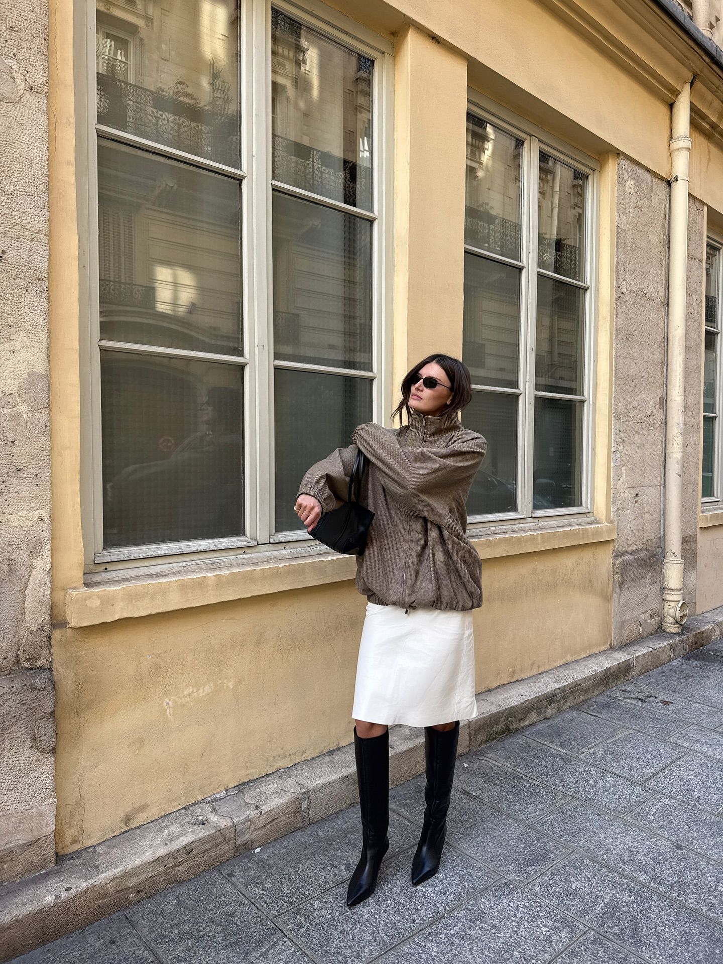 An image of an influencer in Paris wearing a beige windbreaker, white knee-length skirt and black knee-hight boots