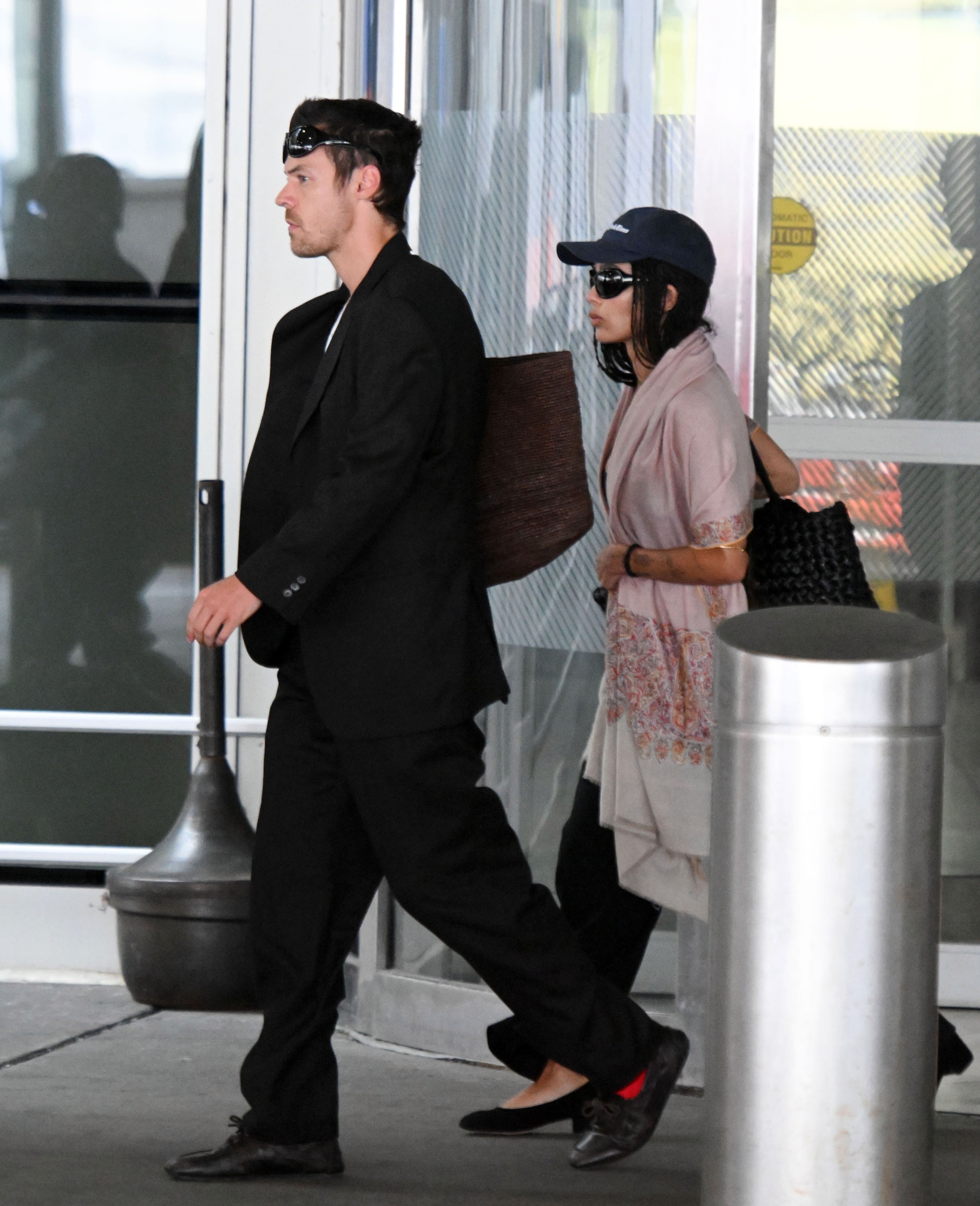 An image of Zoe Kravitz wearing an oversized shawl, black trousers, black ballet pumps anda woven leather bag at JFK.