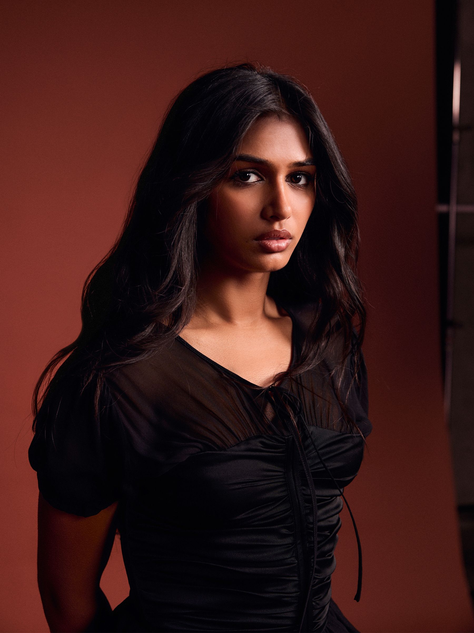 Priyanka Kedia is photographed by Kat and Mariel Tyler in front of a rust colored backdrop wearing a white denim jacket, black shorts, and ankle strap heels.