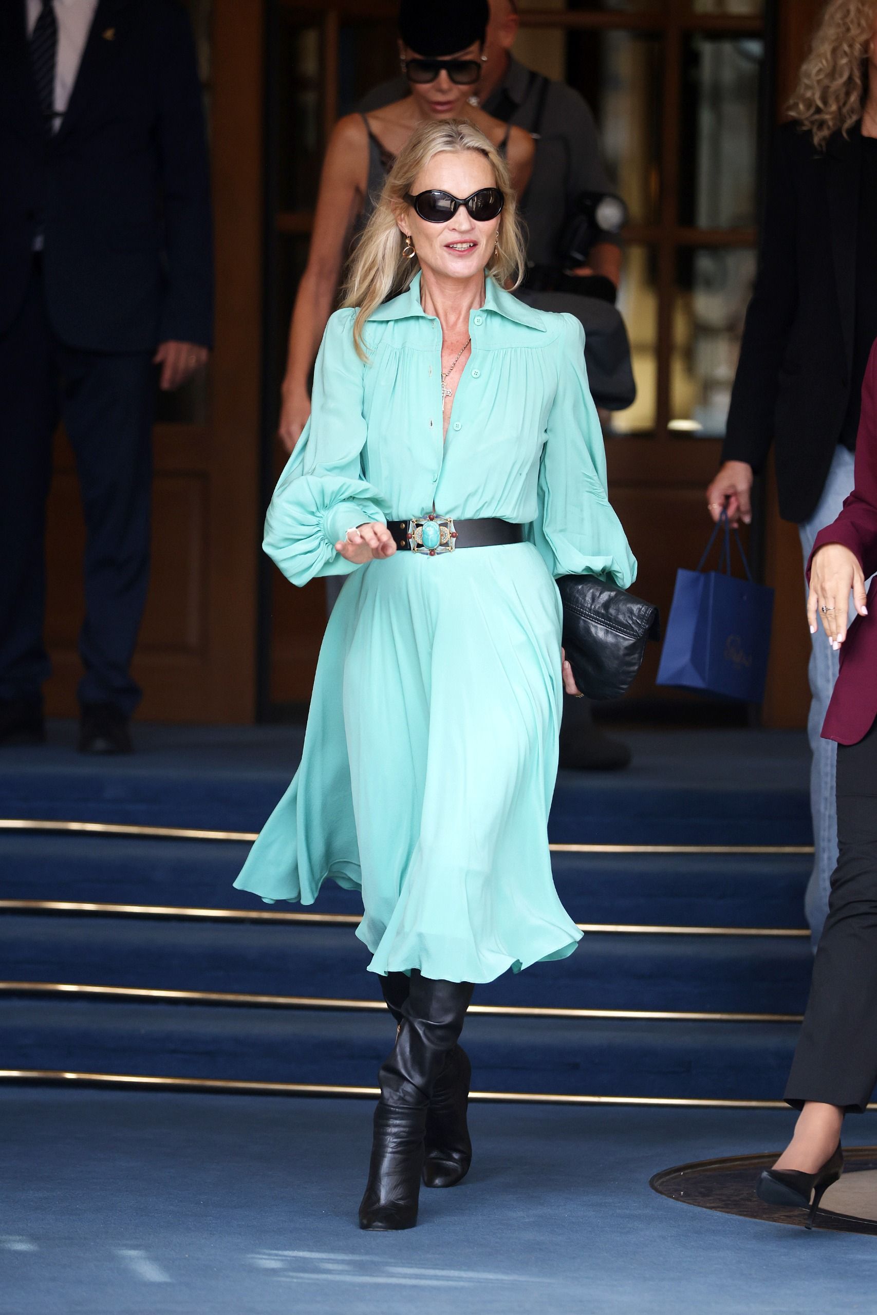 Kate Moss steps outside the Ritz Hotel in Paris during Paris Fashion Week. She wears a turquoise dress with black knee-high boots and a black leather belt.
