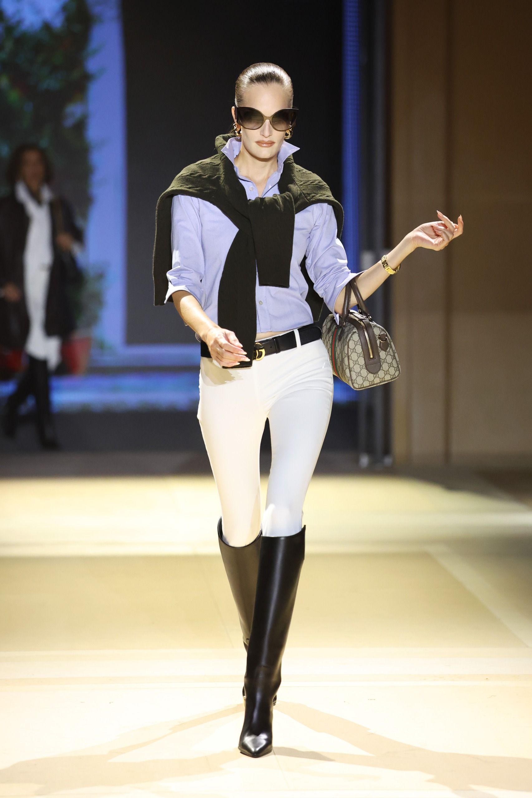 Model walks the Gucci SS27 Runway wearing jodhpur leggings tucked into a shirt with a jumper over the shoulder and black knee-high boots.