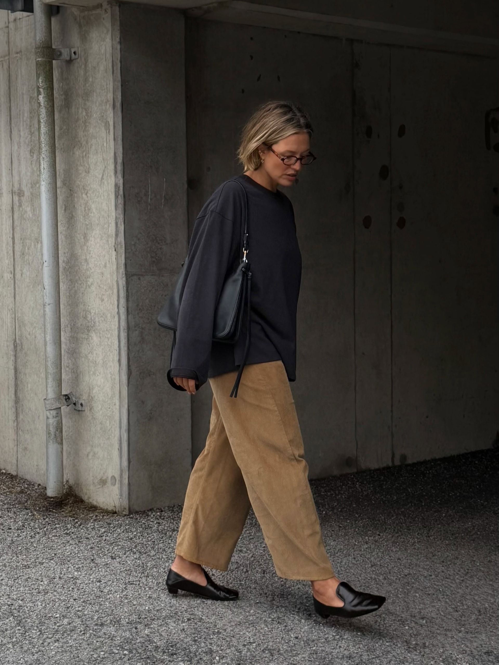 An image of an influencer wearing an autumn shoe trend, heeled loafers, with beige trousers and a navy long-sleeve top carrying a black leather handbag