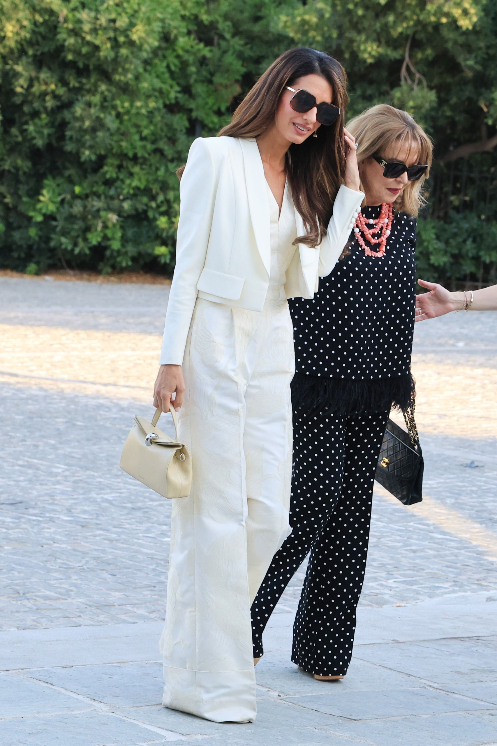 Amal Clooney wears a cropped white blazer and wide-leg pants outfit, while her mom wears a black top and matching trousers with white polka dots.