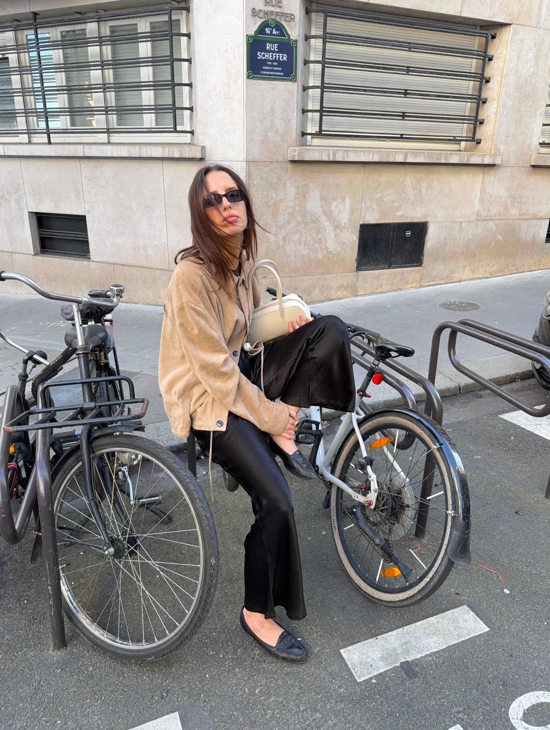 An image of an influencer in Paris holding a white baguette bag and wearing a light beige suede jacket, black satin trousers and black loafers sitting on a bike rack