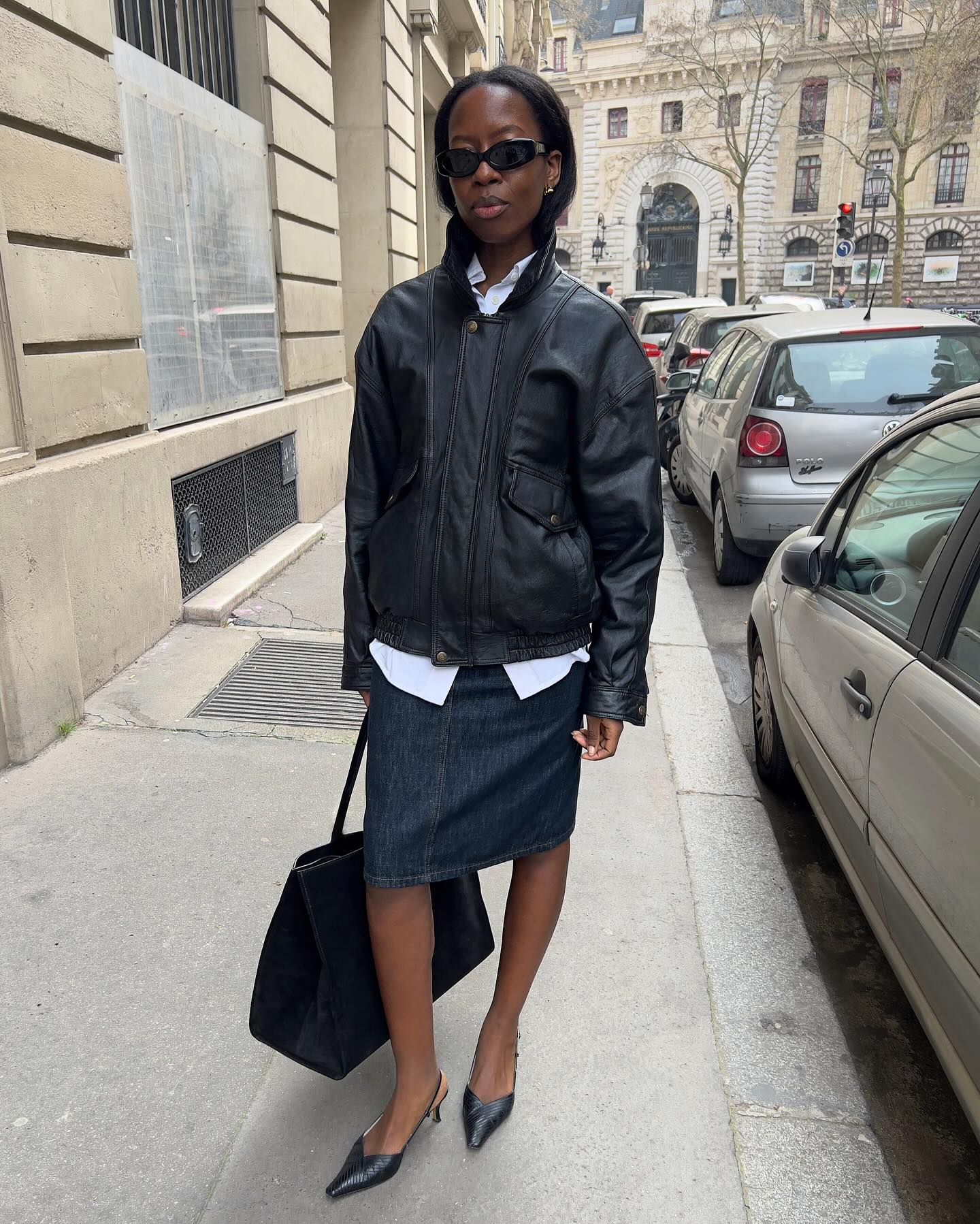 An image of an influencer in Paris wearing sunglasses, a black leather jacket over a white button-down shirt, a midi denim skirt, black kitten heels and holding a suede bowler bag