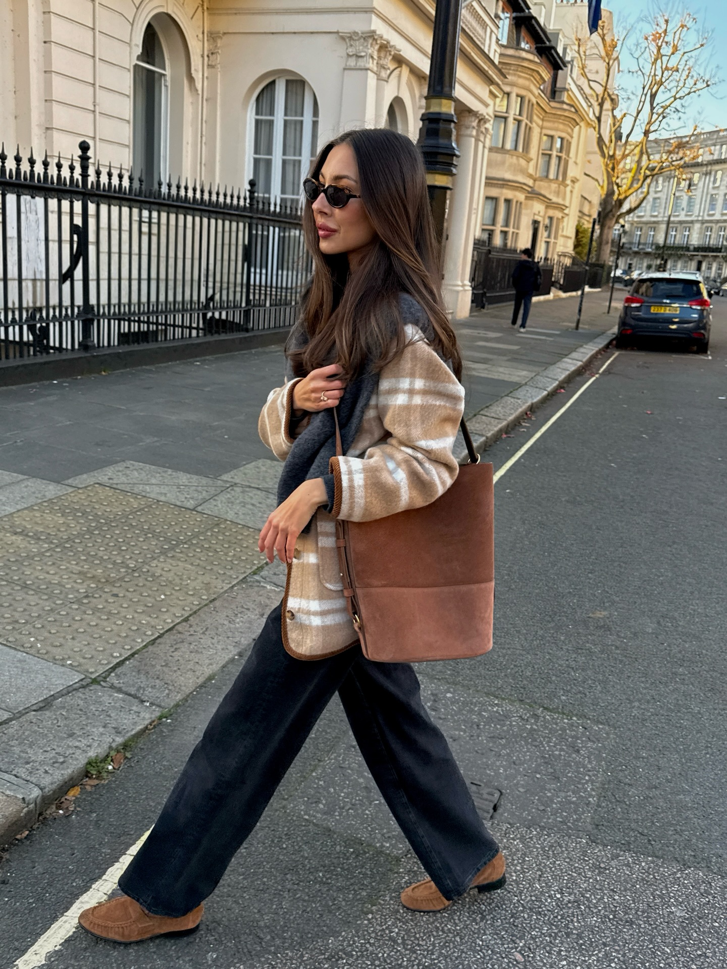 An image of an influencer wearing a plaid beige jacket, black jeans and brown gathered loafers carrying a brown suede tote