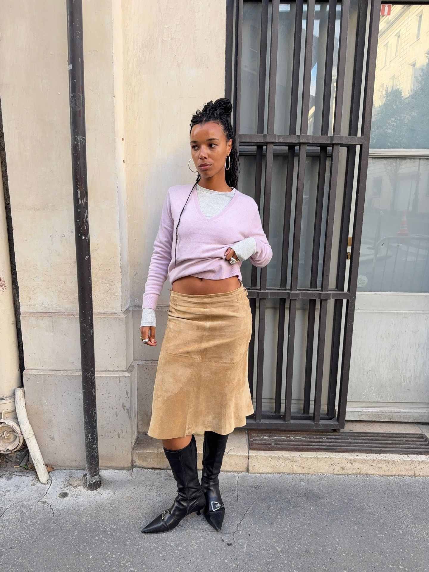 An image of an influencer wearing a light pink knit over a light grey long-sleeve top with a beige suede midi skirt and black kitten heel knee-high boots