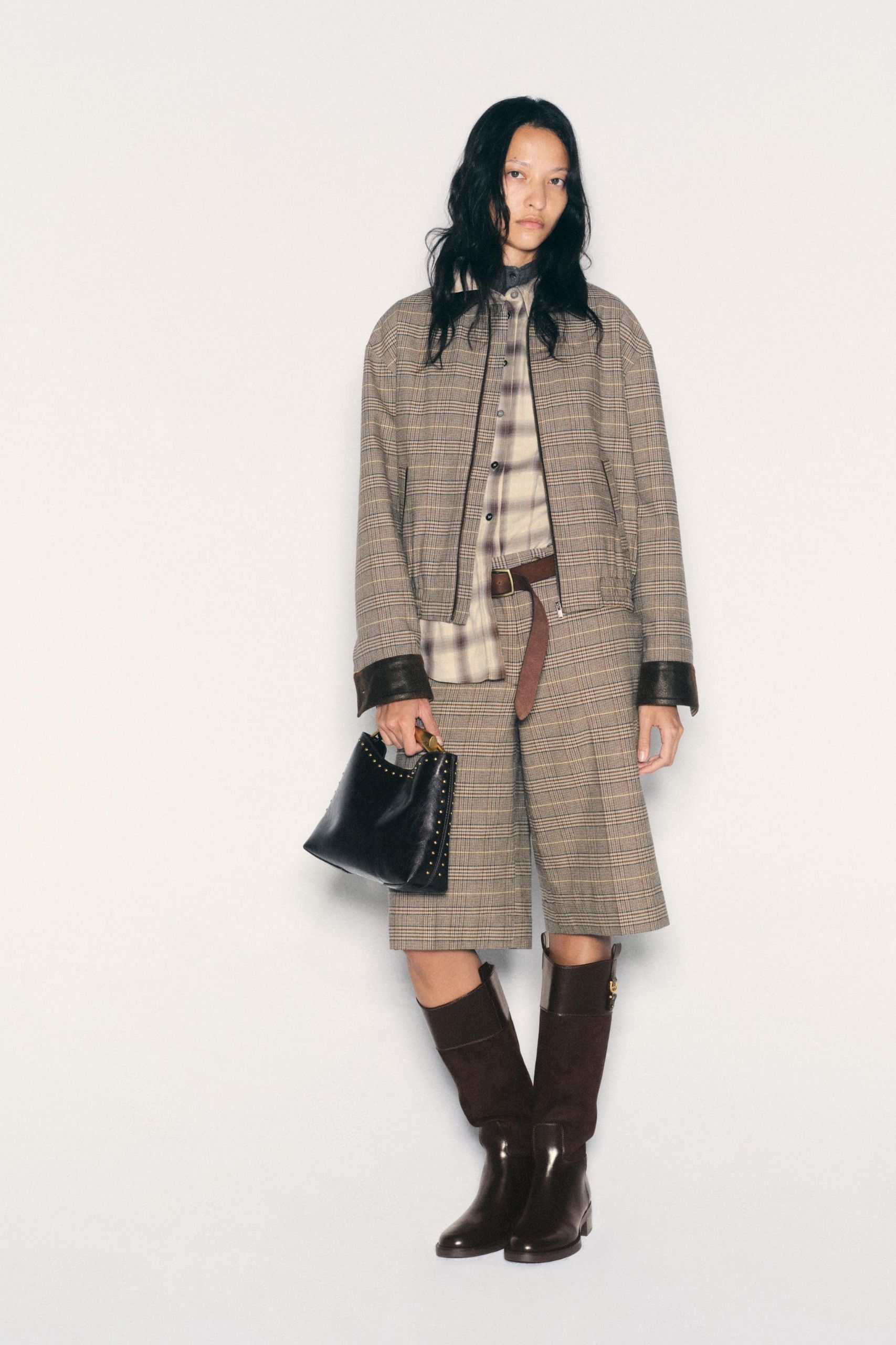 Woman wears matching bomber and Bermuda trouser short set with a plaid shirt and black patent boots.