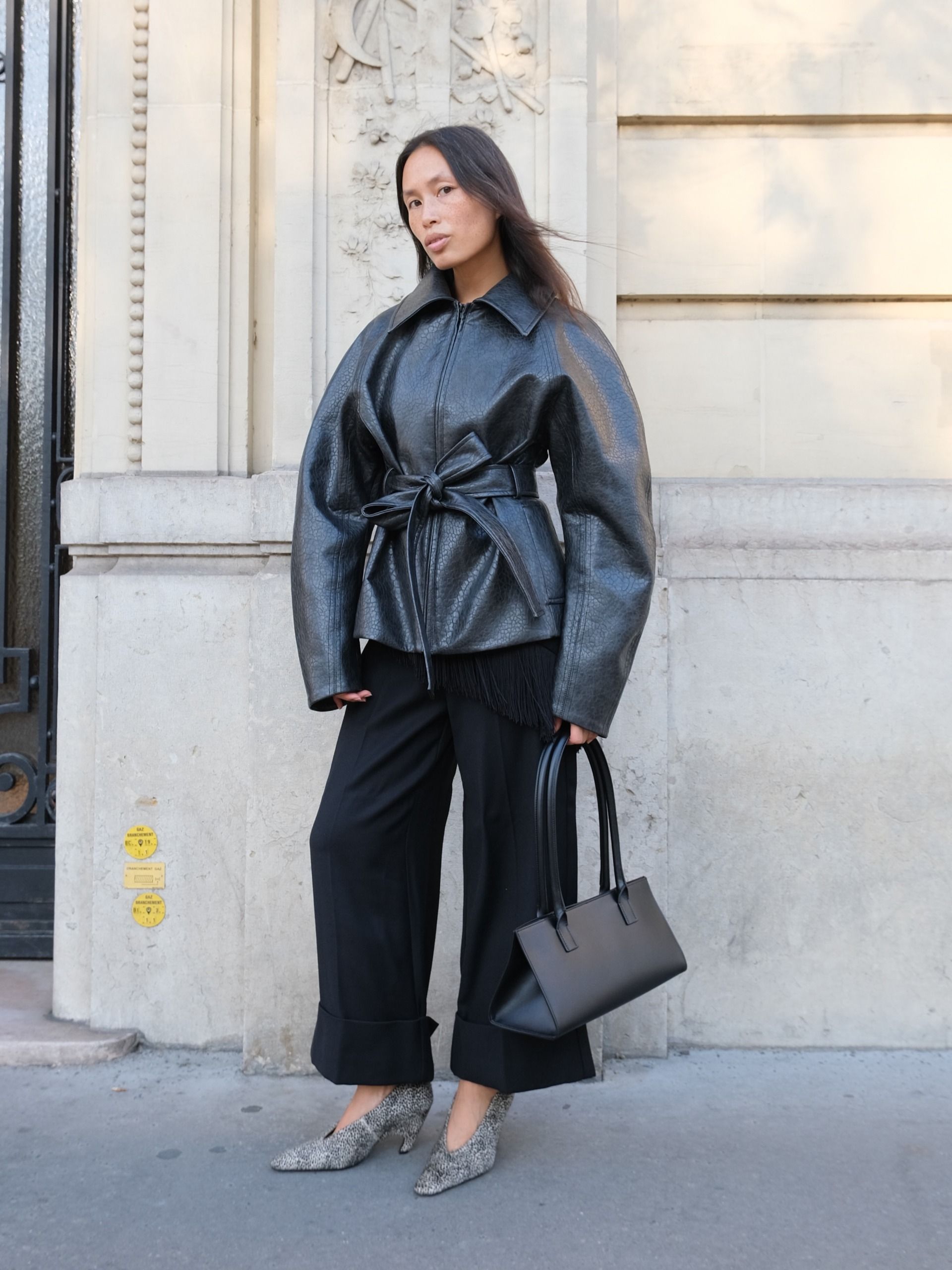 An image of an influencer in Paris wearing a leather waisted jacket with black wide-leg trousers and snake-print heels holding a black handbag