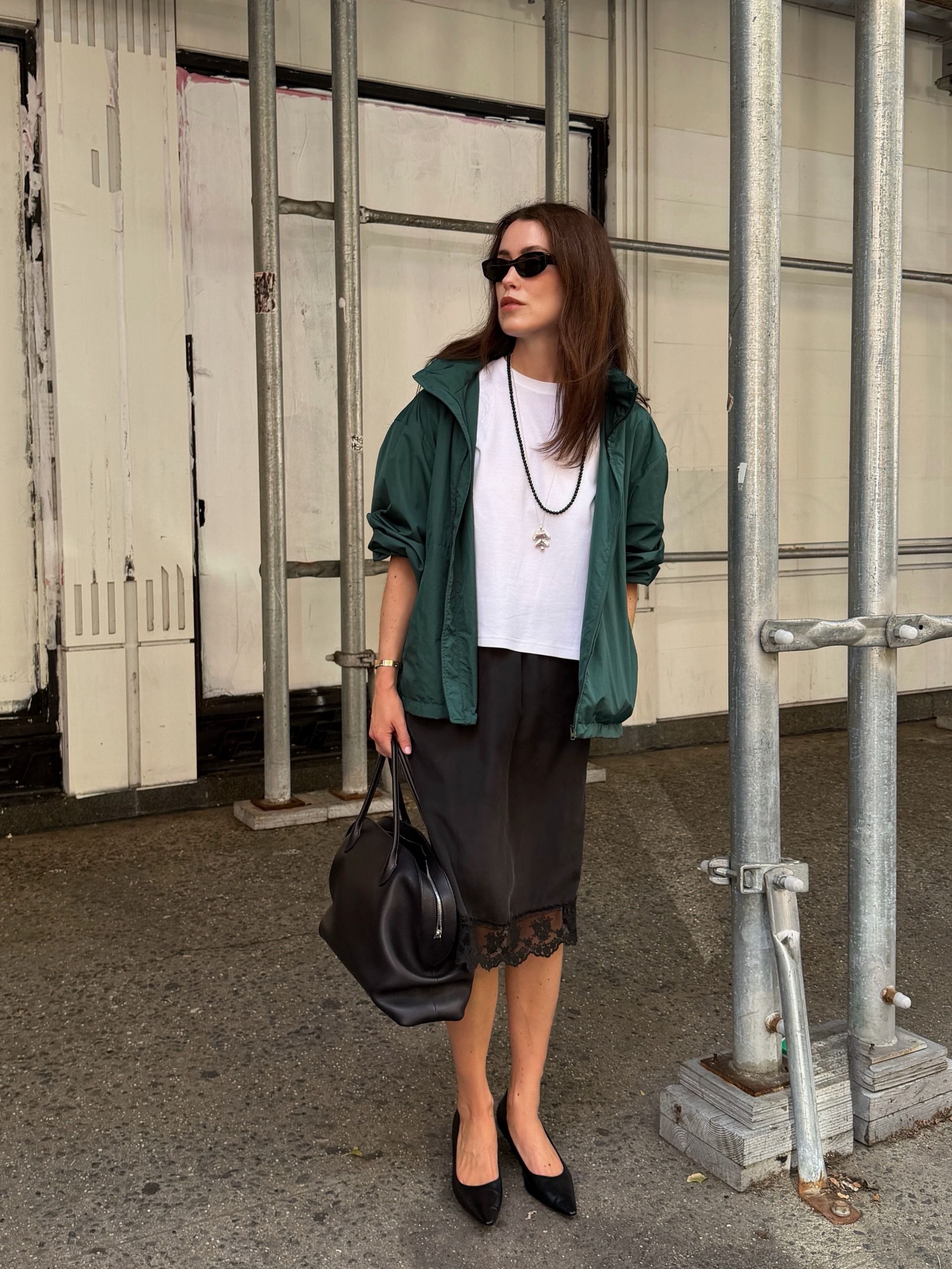 fashion influencer wearing a green windbreaker, white T-shirt, and lace-trim black knee skirt with a The Row Agnes bag