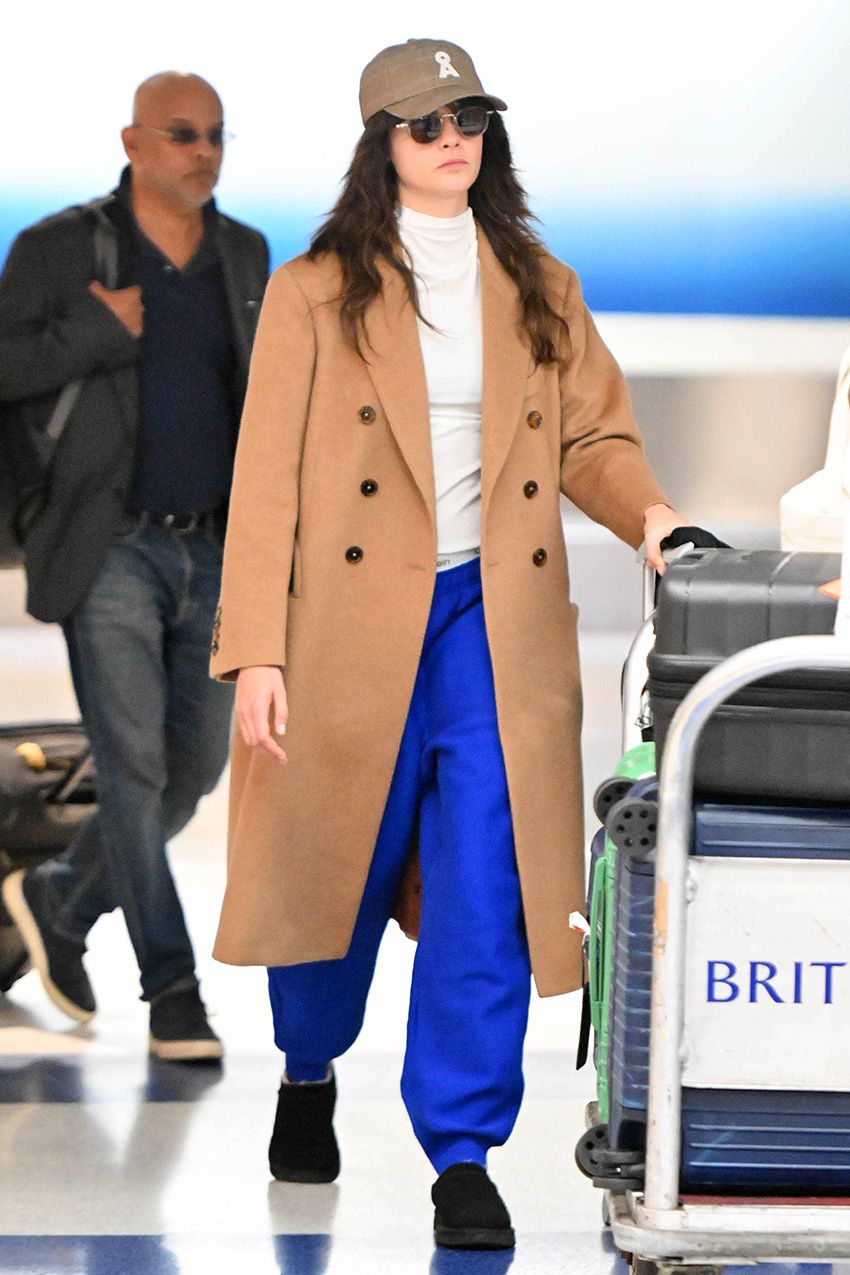 Cara Delevingne wearing cobalt blue pants with a camel coat, cream turtleneck and black suede mules at JFK airport