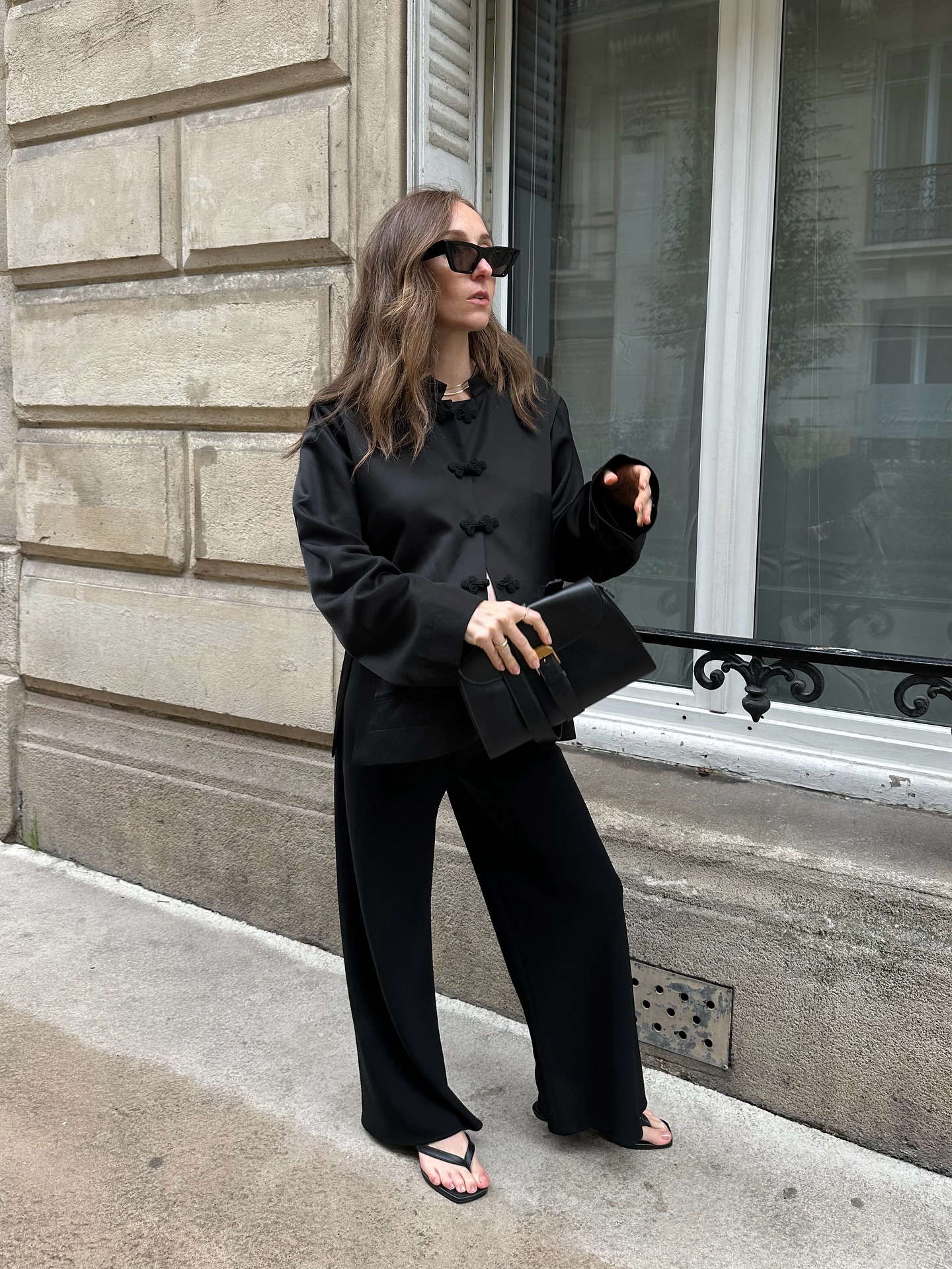 An image of an influencer in Paris wearing a black frog-fastening jacket, black trousers, black flip-flops and holding a black leather clutch